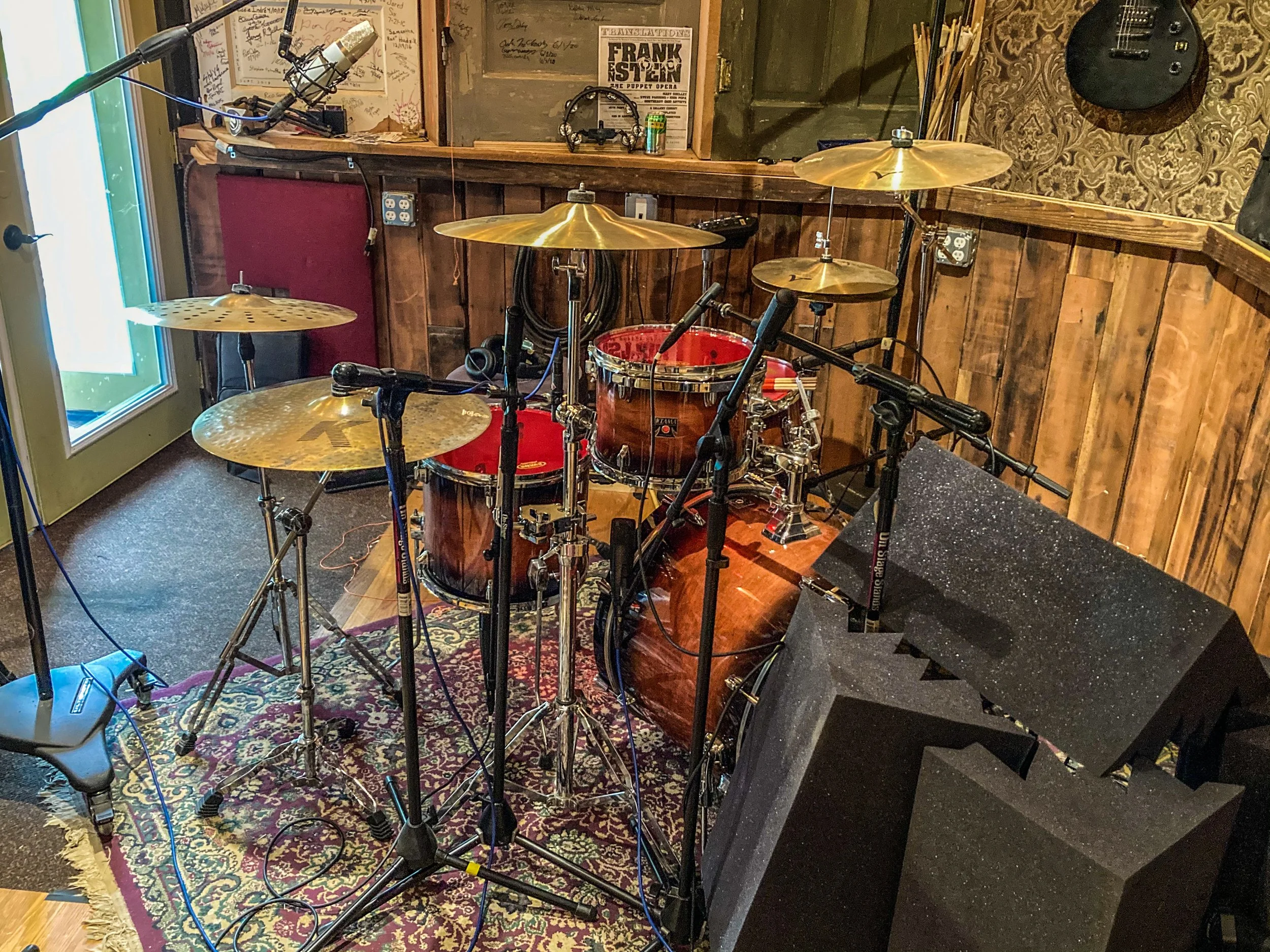 A drum set in a recording studio with microphones, cymbals, drums, and speakers, set up on a patterned rug against wooden panel walls.