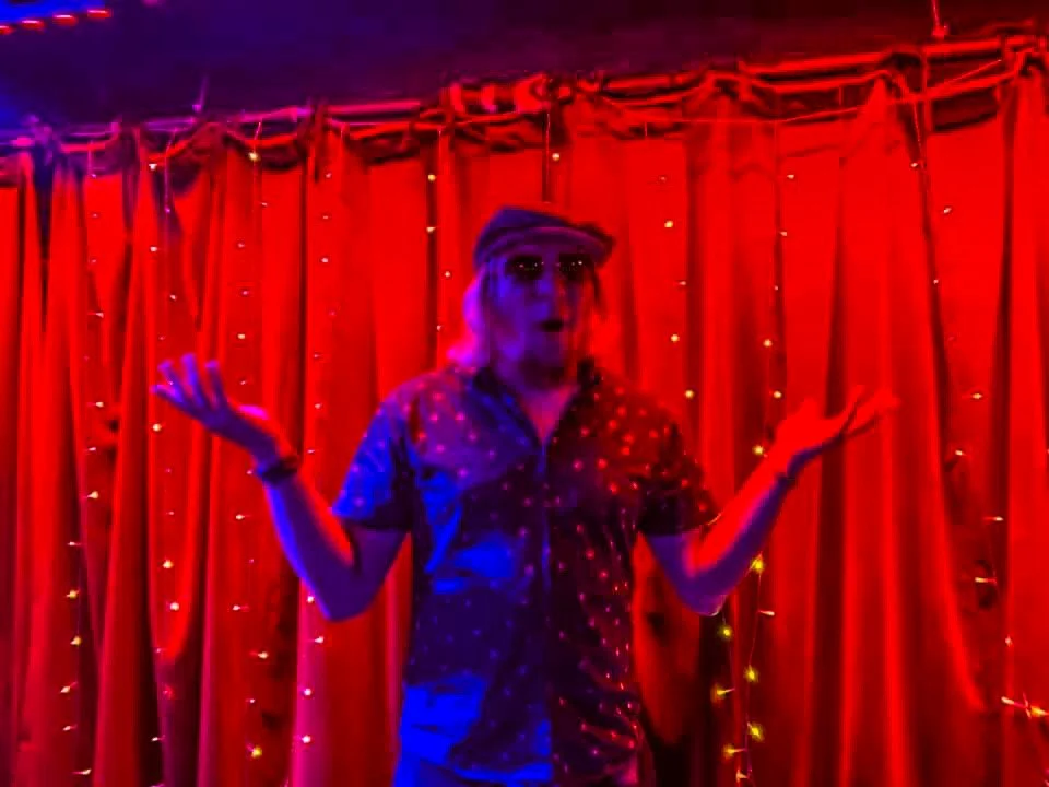 Person on stage in front of red curtain with lights, wearing sunglasses and a patterned shirt, gesturing with their hands.