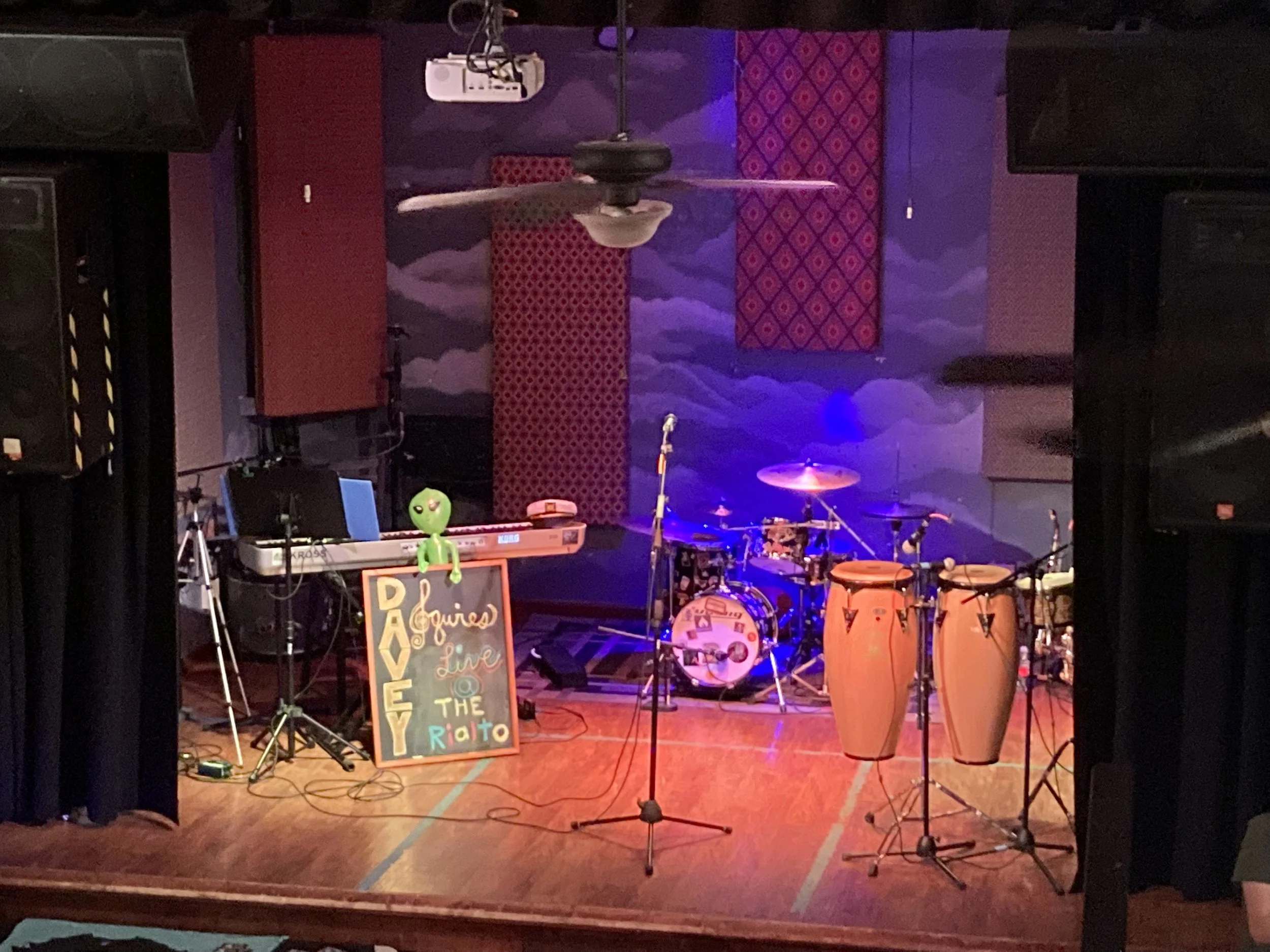 Empty stage with musical instruments and decorations, including drums, congas, and a keyboard, with a sign that reads 'A Guitars Live at the Rialto' and a green alien plush on the sign. The background has colorful murals and wall hangings.