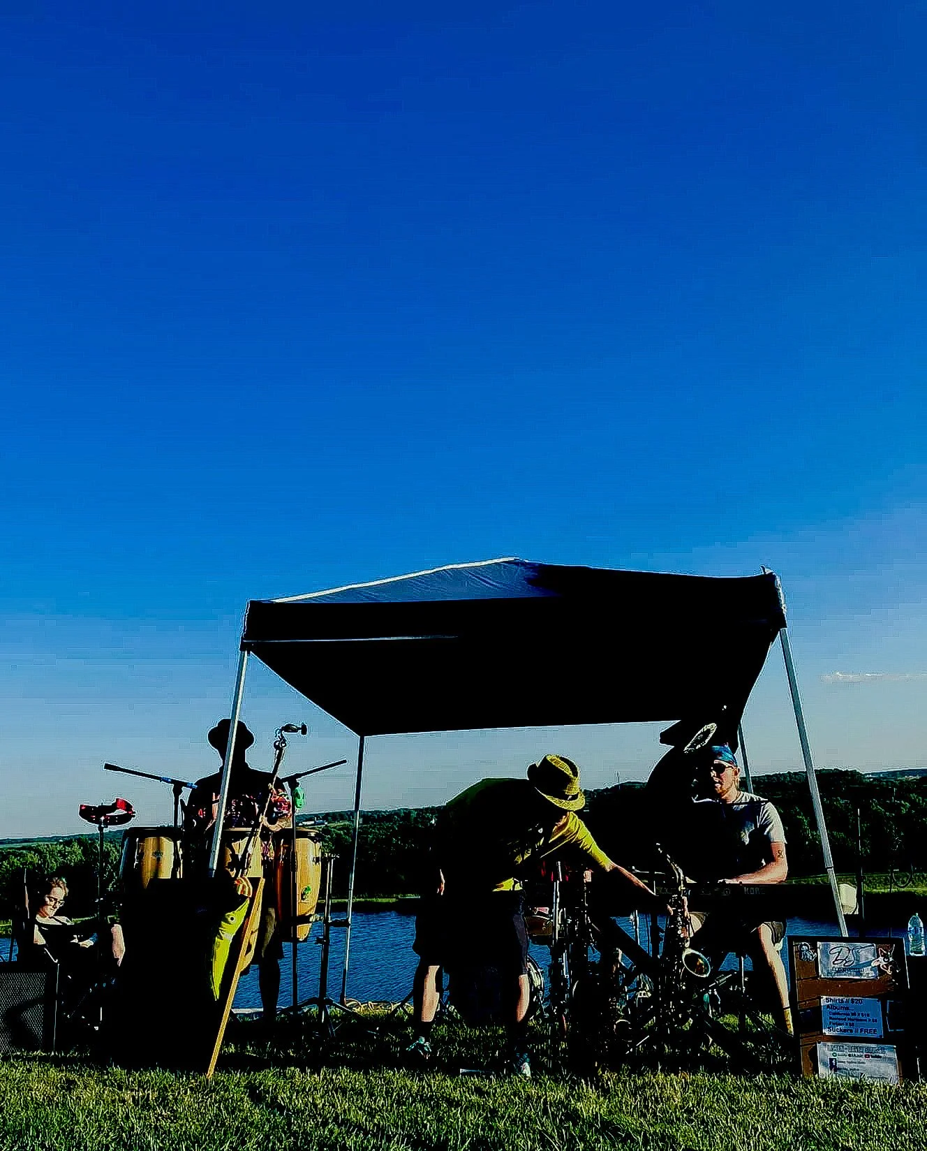 People under a canopy near a body of water, with musical instruments and a sign displayed on the grass, during sunny weather.