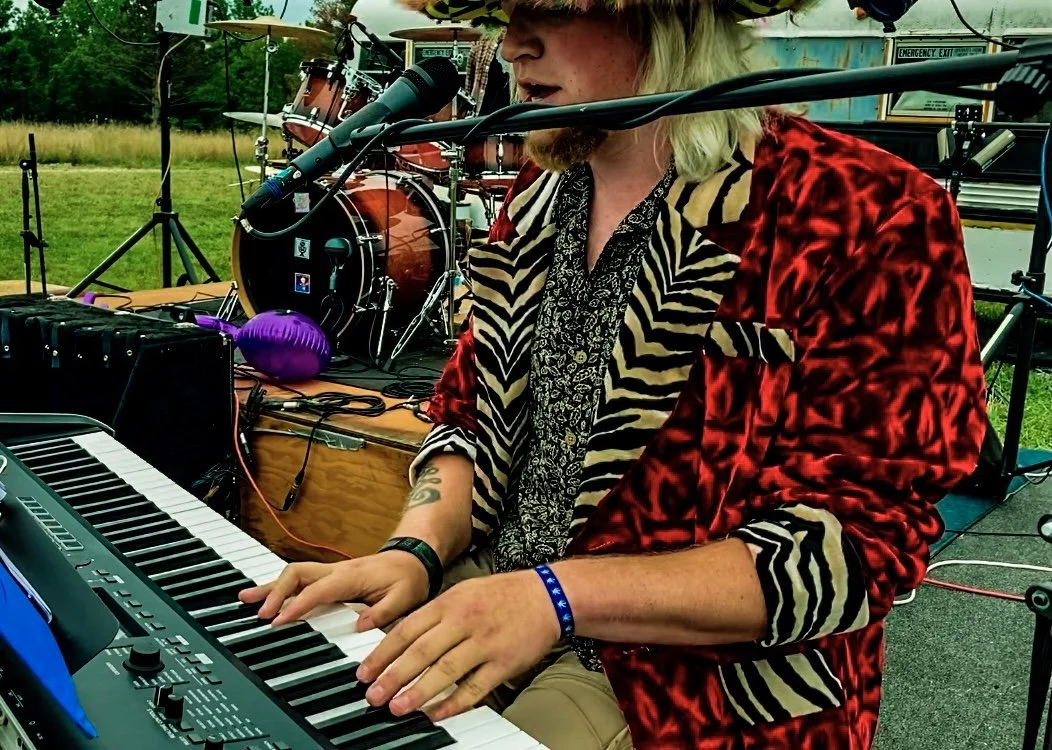 A man playing a keyboard outdoors during a live music performance, with drums set up in the background.