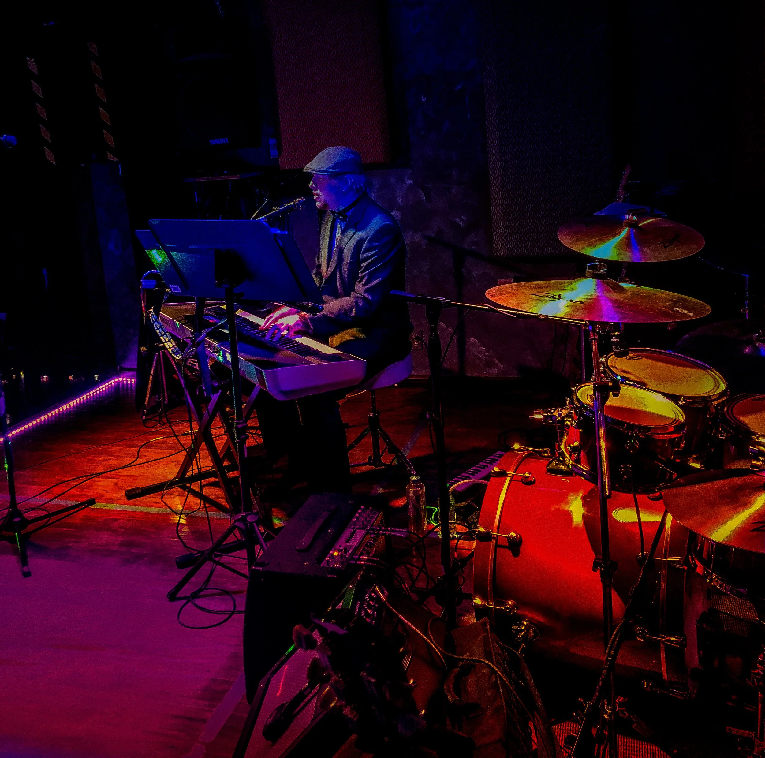 A musician playing a keyboard on stage with drums behind them, illuminated by colorful stage lights.