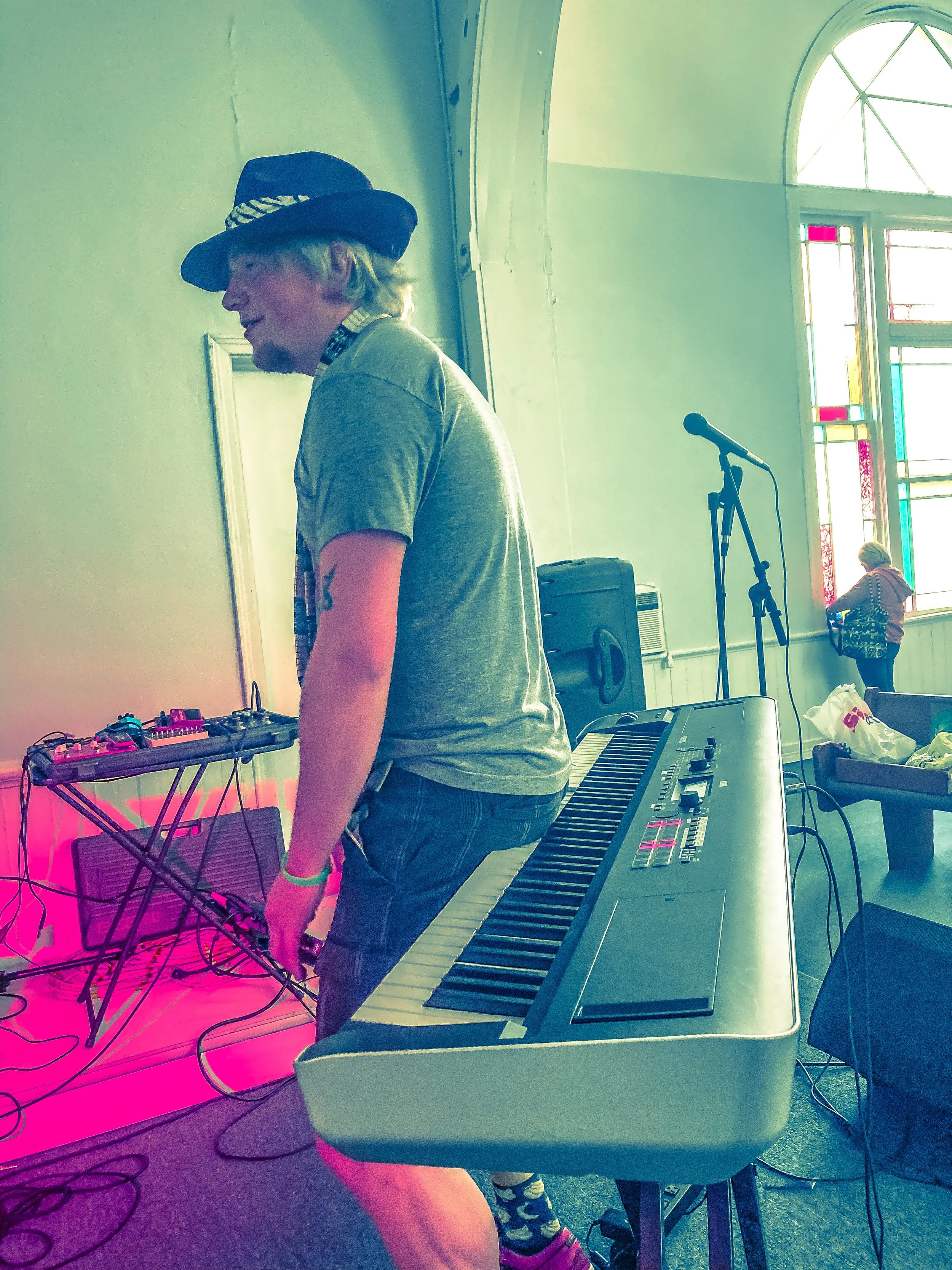 Man wearing hat and gray t-shirt standing next to a keyboard at a music setup inside a room with high arched windows.