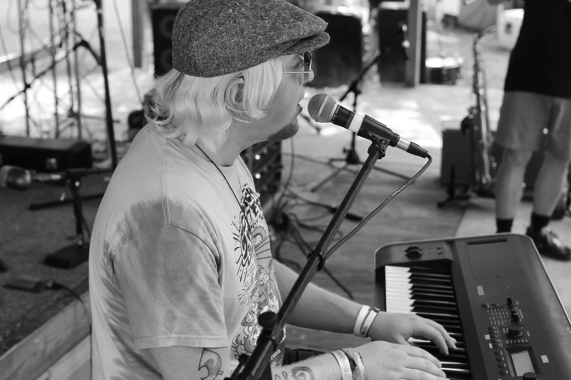 A man with long hair, glasses, and a hat playing a keyboard at an outdoor event, with microphones and audio equipment nearby.