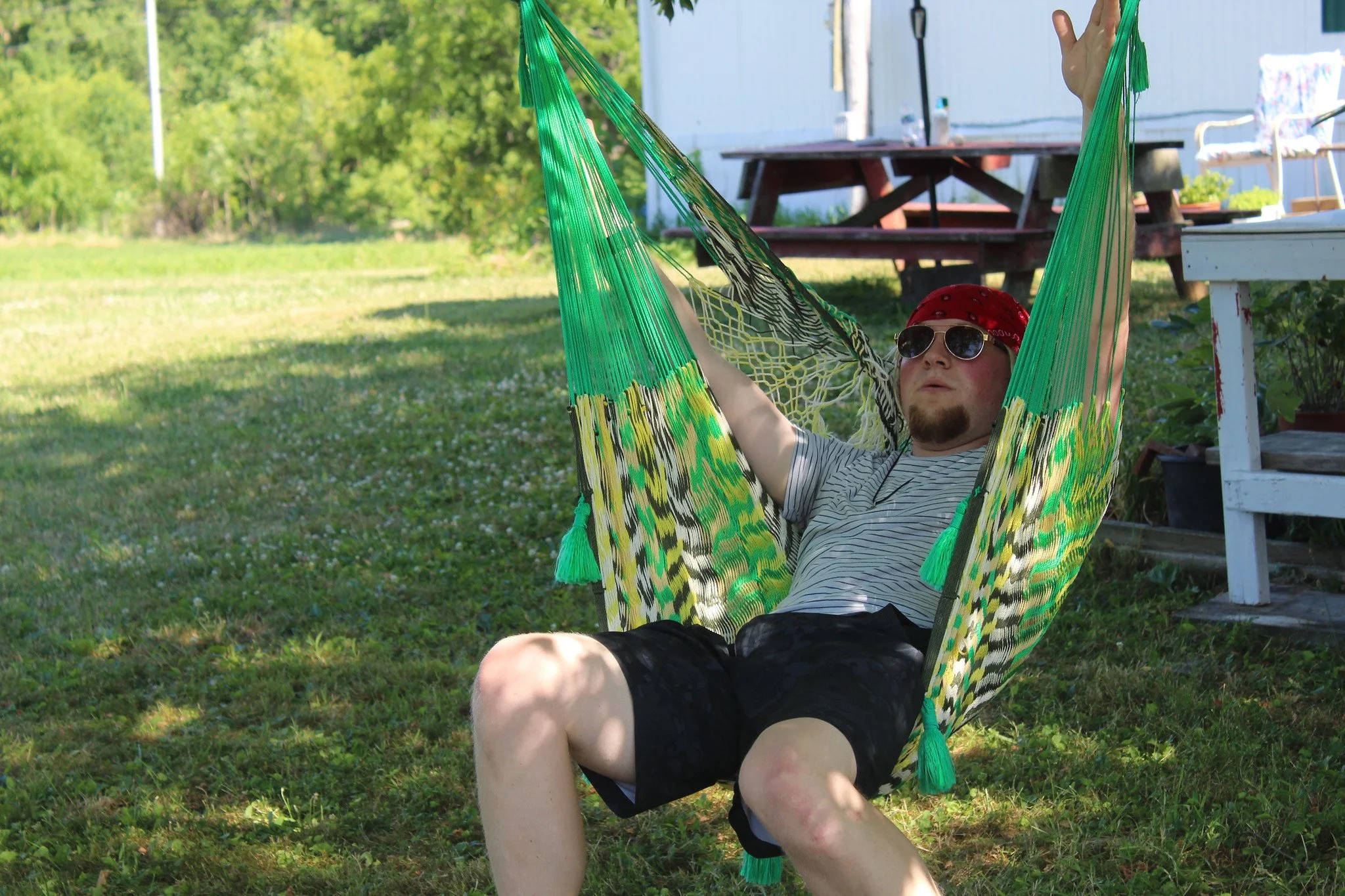 A man relaxing in a hammock outdoors on a sunny day, wearing sunglasses, a red bandana, a striped t-shirt, and black shorts.