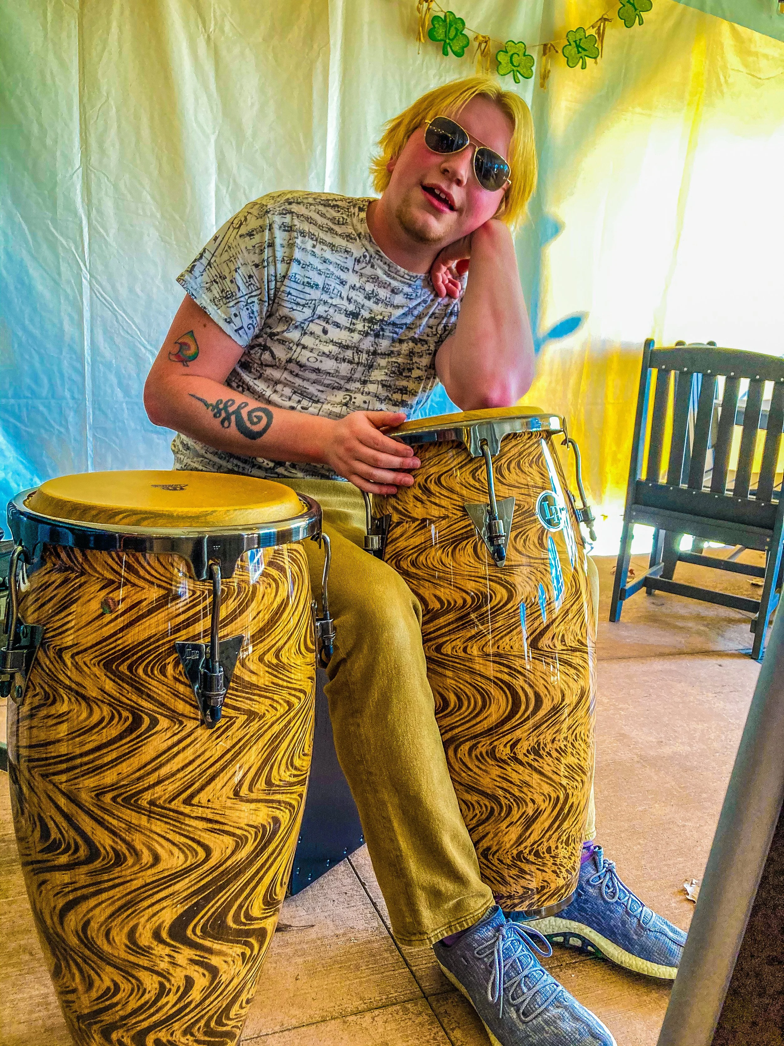 A person sitting behind two large yellow and black marbled drums, wearing sunglasses, a graphic t-shirt, khaki pants, and sneakers, with a festive backdrop of green shamrocks hanging in the background.