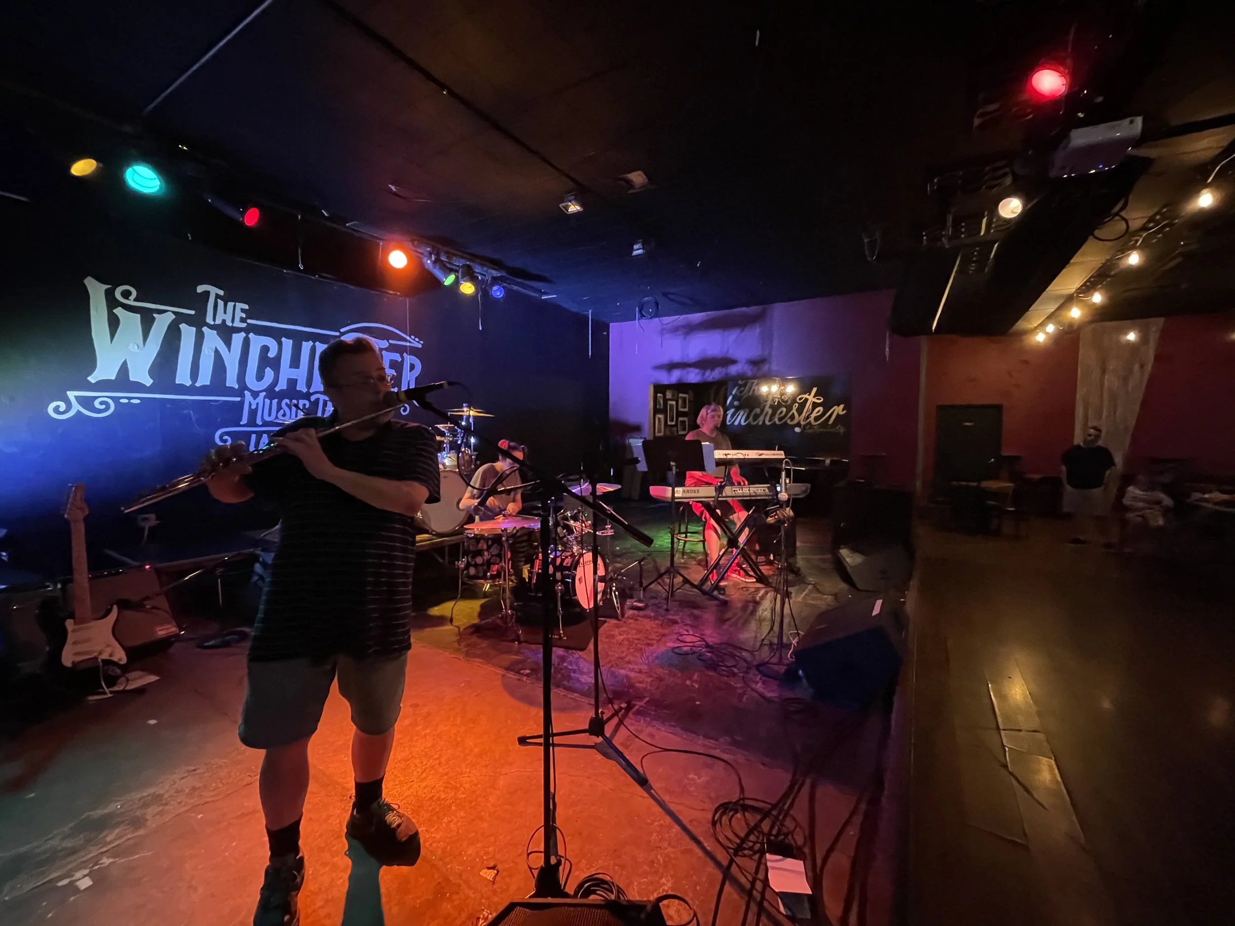 Band performing on stage at Winchester Music Tavern, with band members playing flute, drums, and keyboard, colorful stage lights, and audience seated in the background.