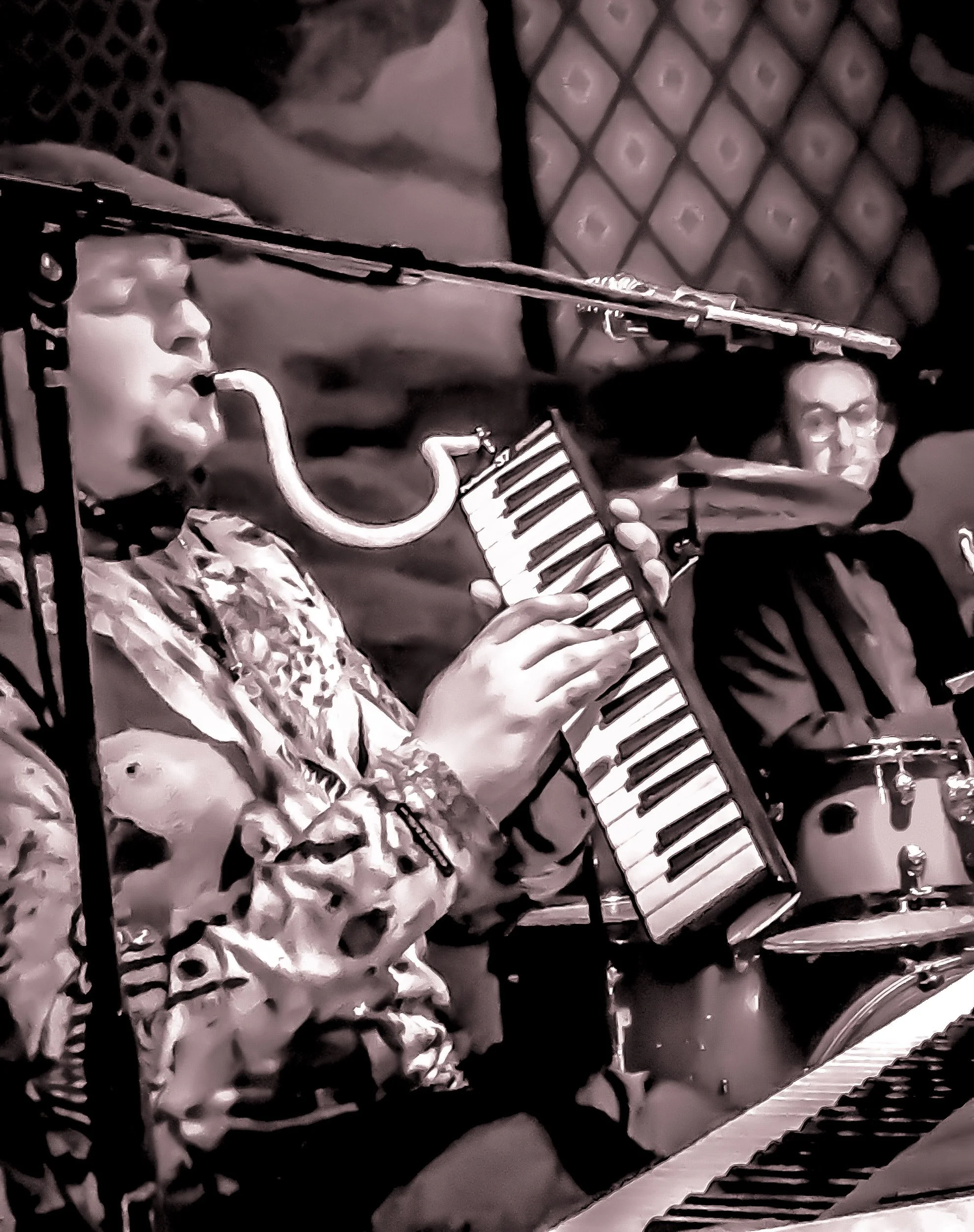 Jazz musicians performing, with one playing a saxophone and a small keyboard, and another on drums, in a dimly lit venue.