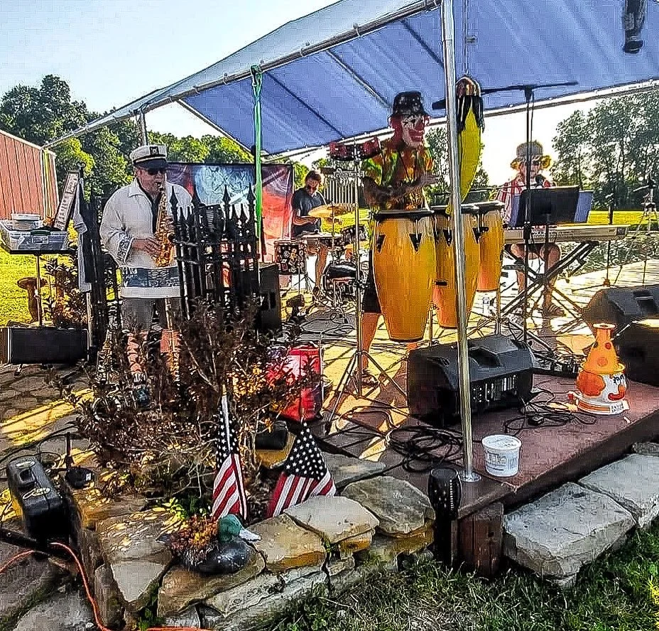 A live outdoor band performing under a tent during the daytime, with musical instruments including drums, congas, keyboard, and saxophone, surrounded by flags and decorations, and a rural landscape in the background.