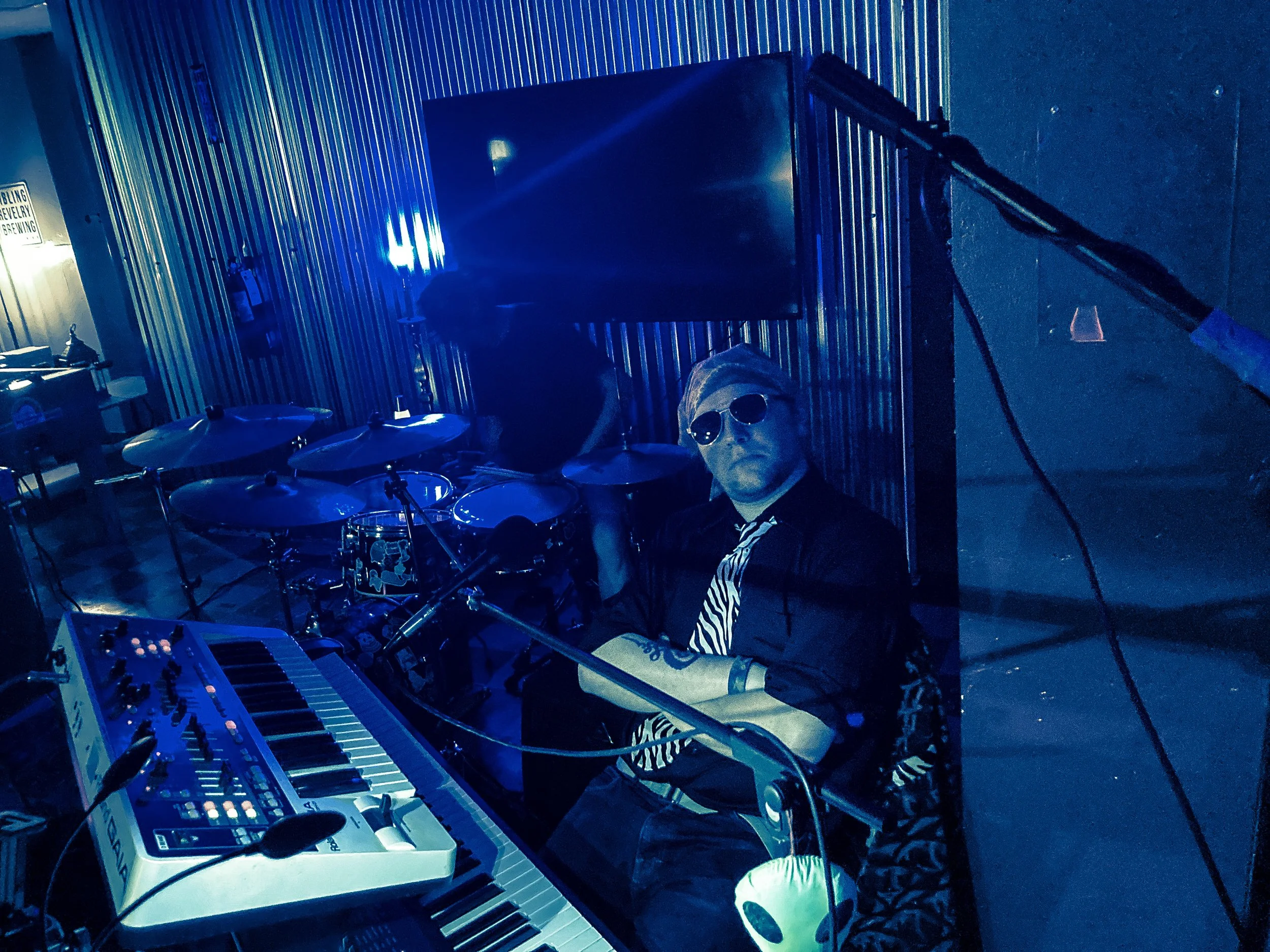 Musician wearing sunglasses seated behind a keyboard, with drums in the background, in a dimly lit setting with blue lighting.