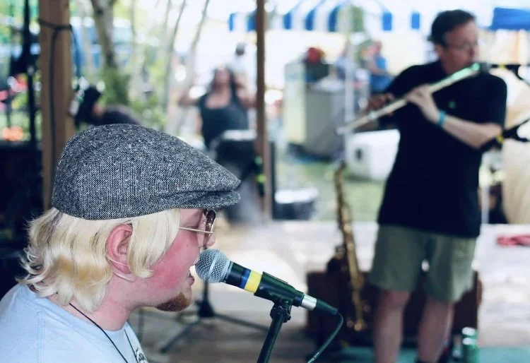 A musician with long blonde hair, wearing a gray flat cap and sunglasses, singing into a microphone at an outdoor event.