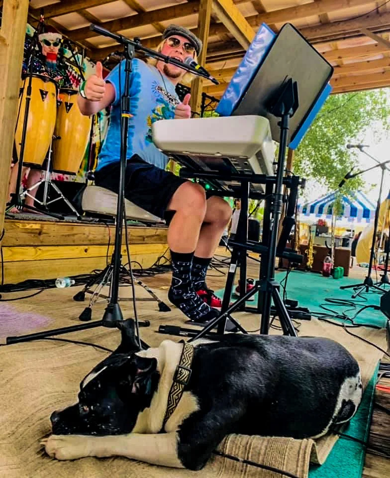 A man wearing sunglasses, a hat, and a blue shirt with a colorful design is sitting at a keyboard, giving a thumbs-up and smiling. There is a microphone in front of him and a dog lying down on a rug in the foreground.