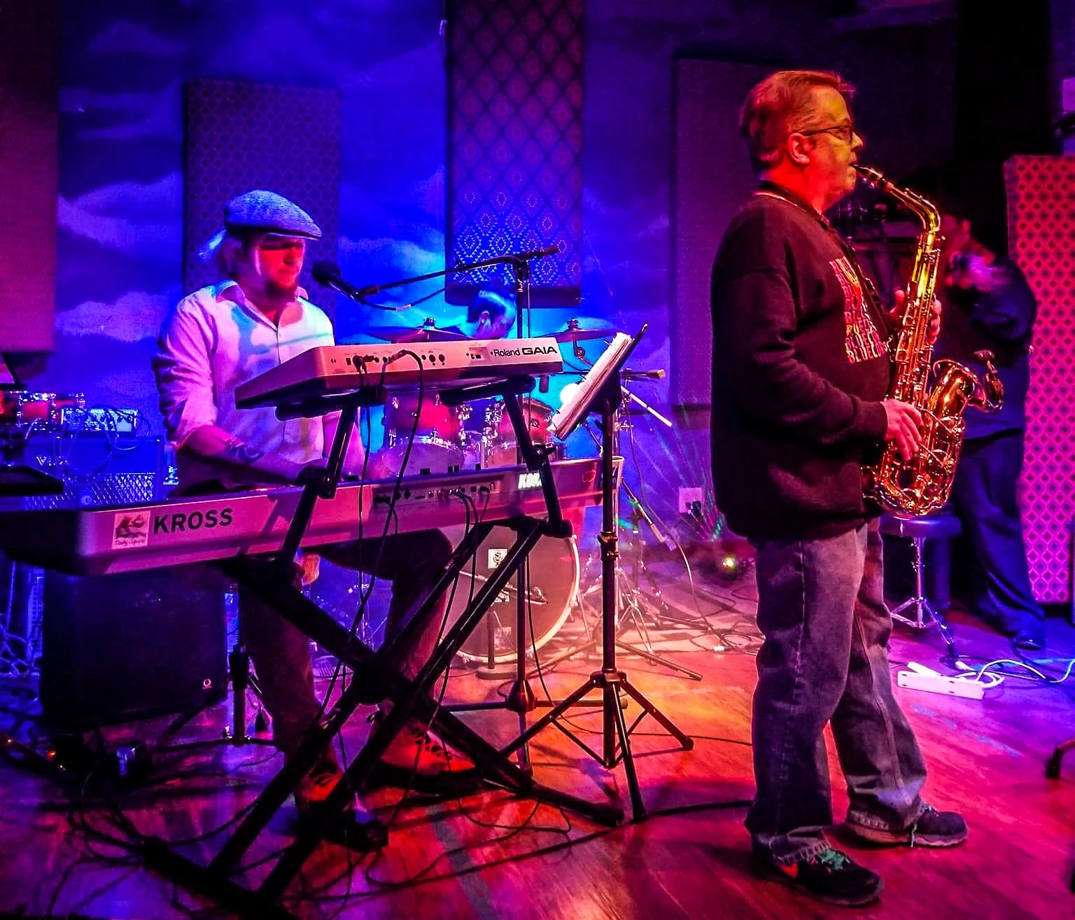 Musicians performing on stage with keyboard, drums, and saxophone in colorful lighting.