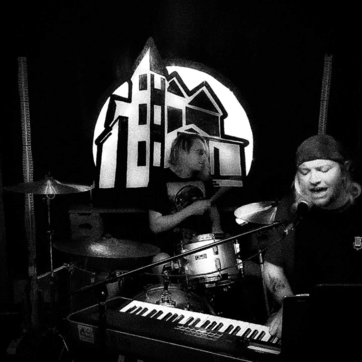 A black and white photo of two people performing music on stage, with a large graphic of a house and moon in the background. One person is singing into a microphone and playing a keyboard, while the other is playing drums.