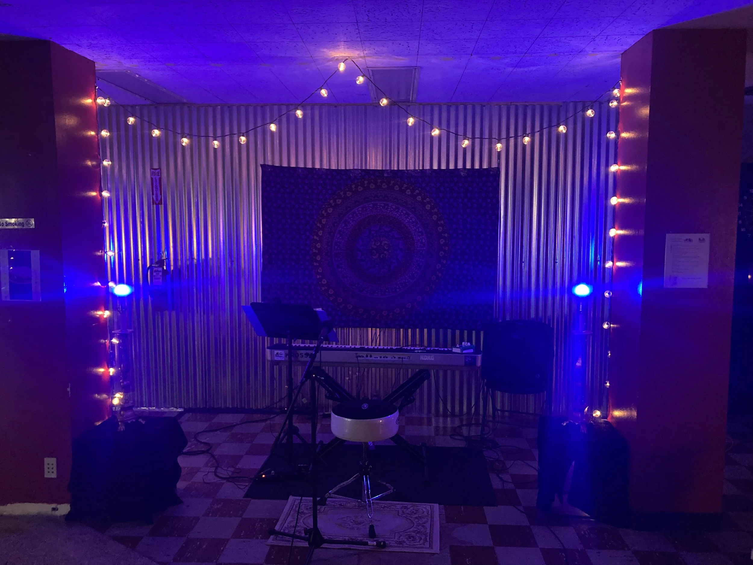 Indoor music setup with microphone, keyboard, and music stand, illuminated by blue and purple lighting, with string lights on the wall.