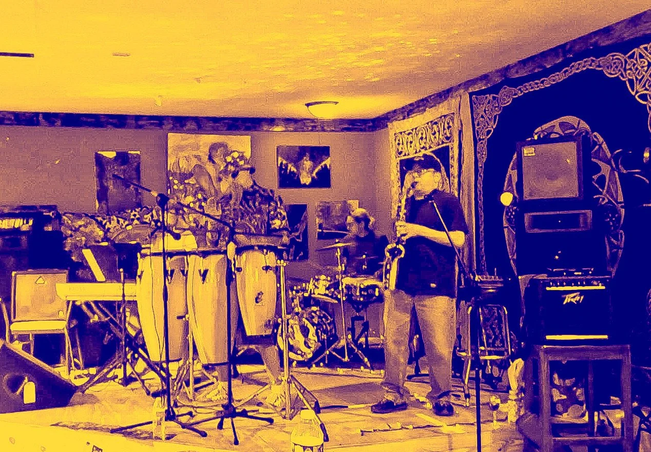 A band performing live on a stage with various musical instruments and microphones, in a room decorated with paintings and a tapestry.