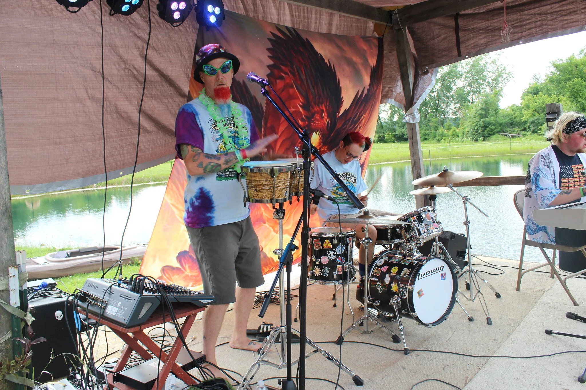 A band performing outdoors near a lake with a large eagle artwork in the background. The band members are dressed casually with colorful accessories, playing drums and other instruments under a canopy.