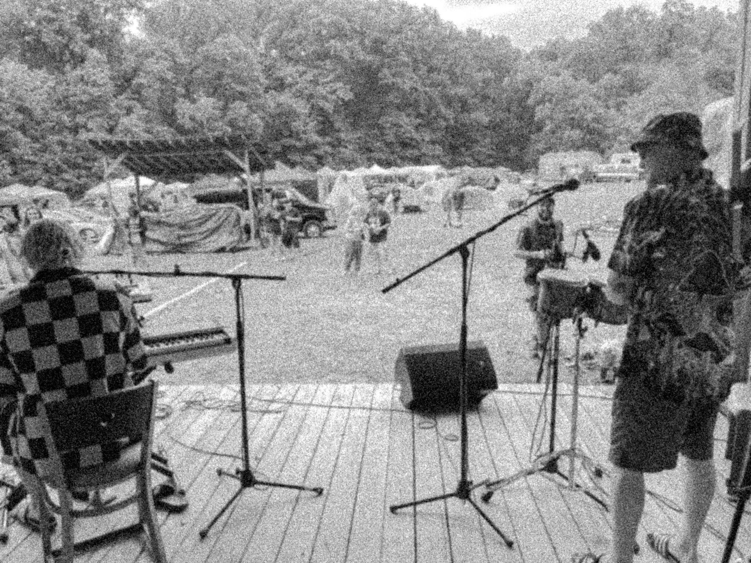 Musicians performing outdoors at a park, with one person singing into a microphone and others playing instruments, surrounded by trees, tents, and parked cars.