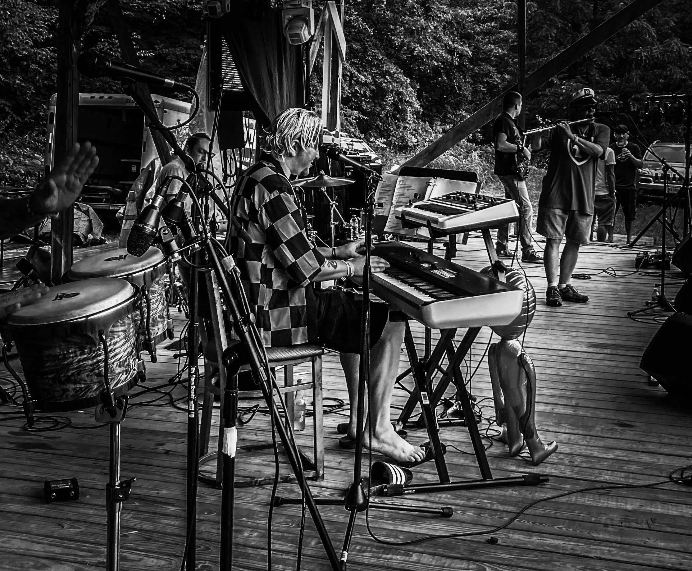 A group of musicians performing on an outdoor wooden stage, with one playing a keyboard and others playing various instruments, surrounded by trees and equipment.