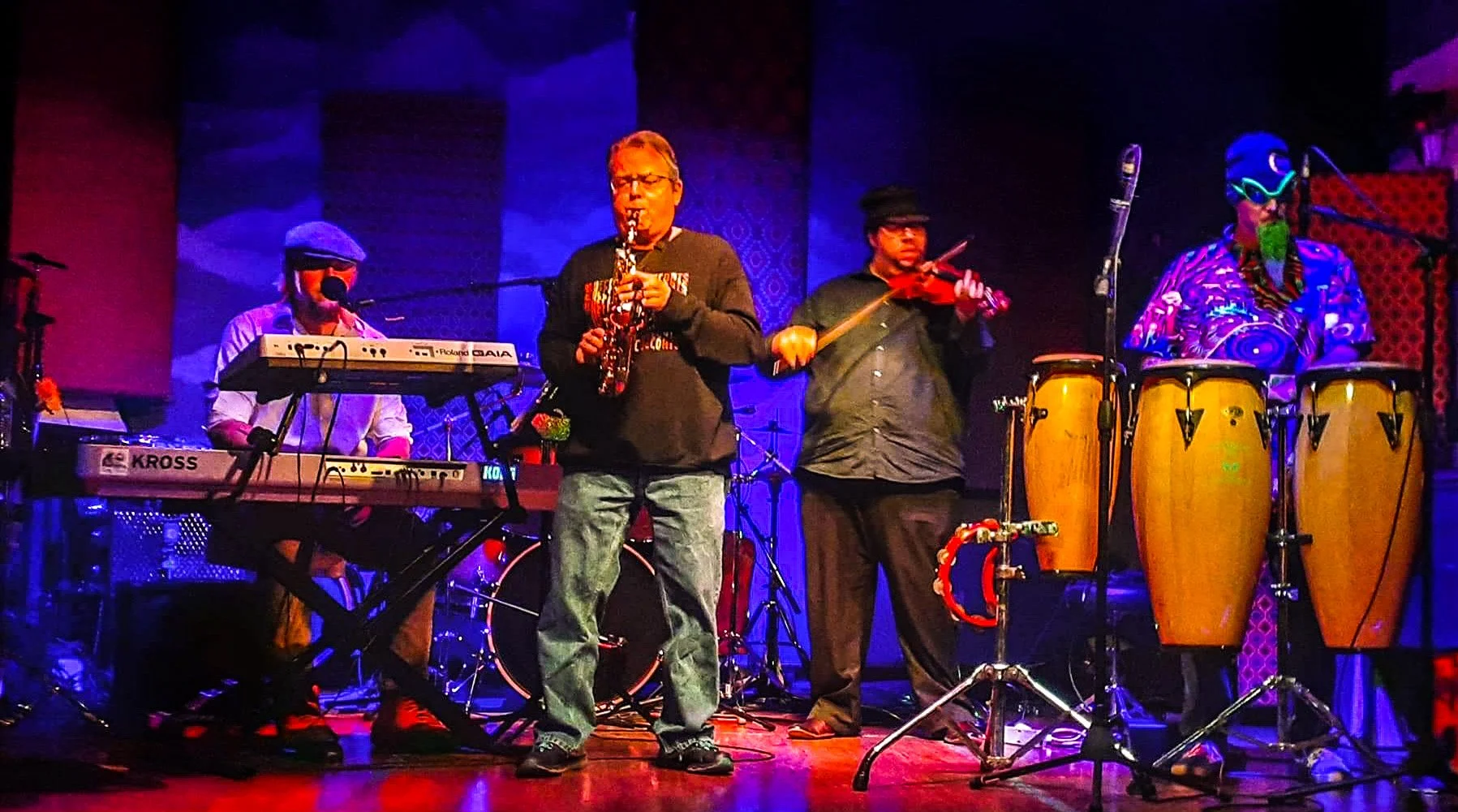 Four musicians performing on stage, with one playing keyboard, one playing saxophone, one playing violin, and one playing conga drums, illuminated by colorful stage lighting.