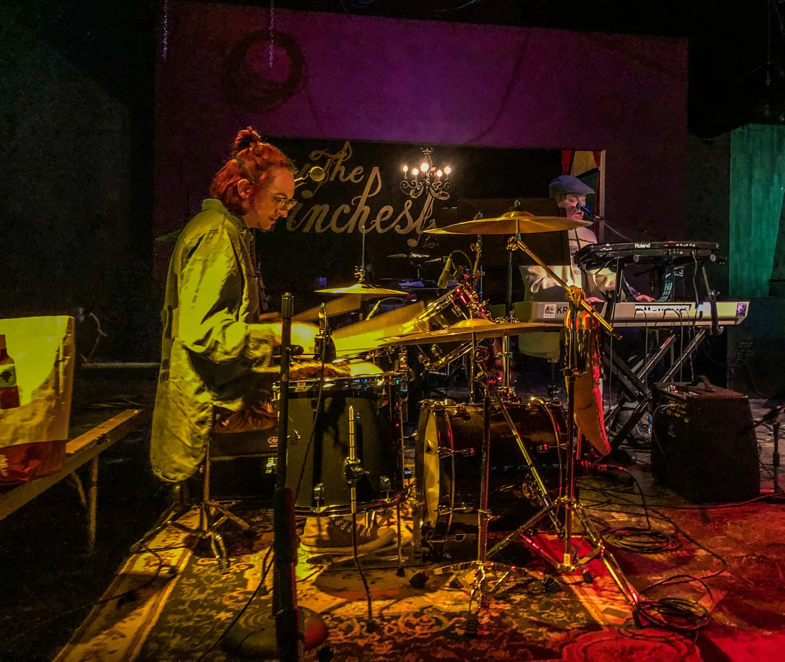 A musical performance at an indoor venue, with a drummer and keyboardist on stage, illuminated by warm lighting. The backdrop features a black banner with gold script. The stage is decorated with a patterned rug and various musical equipment.