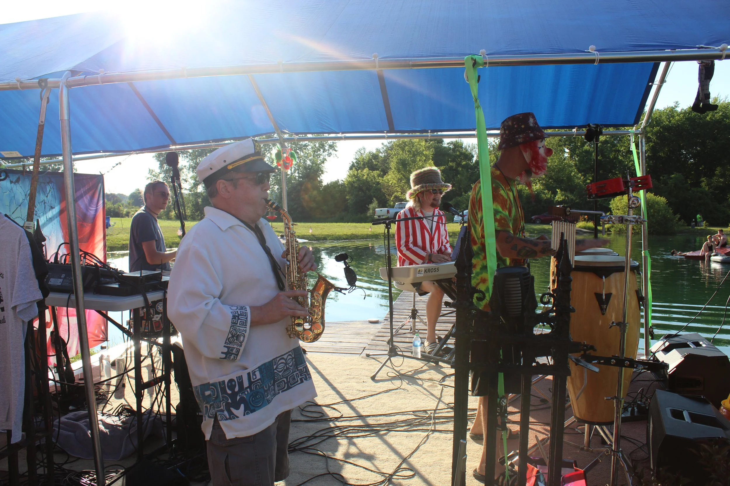 A band performing outdoors under a blue canopy near a lake, with musicians playing instruments including saxophone and drums, and a person on a keyboard wearing a colorful tie-dye shirt and clown makeup.