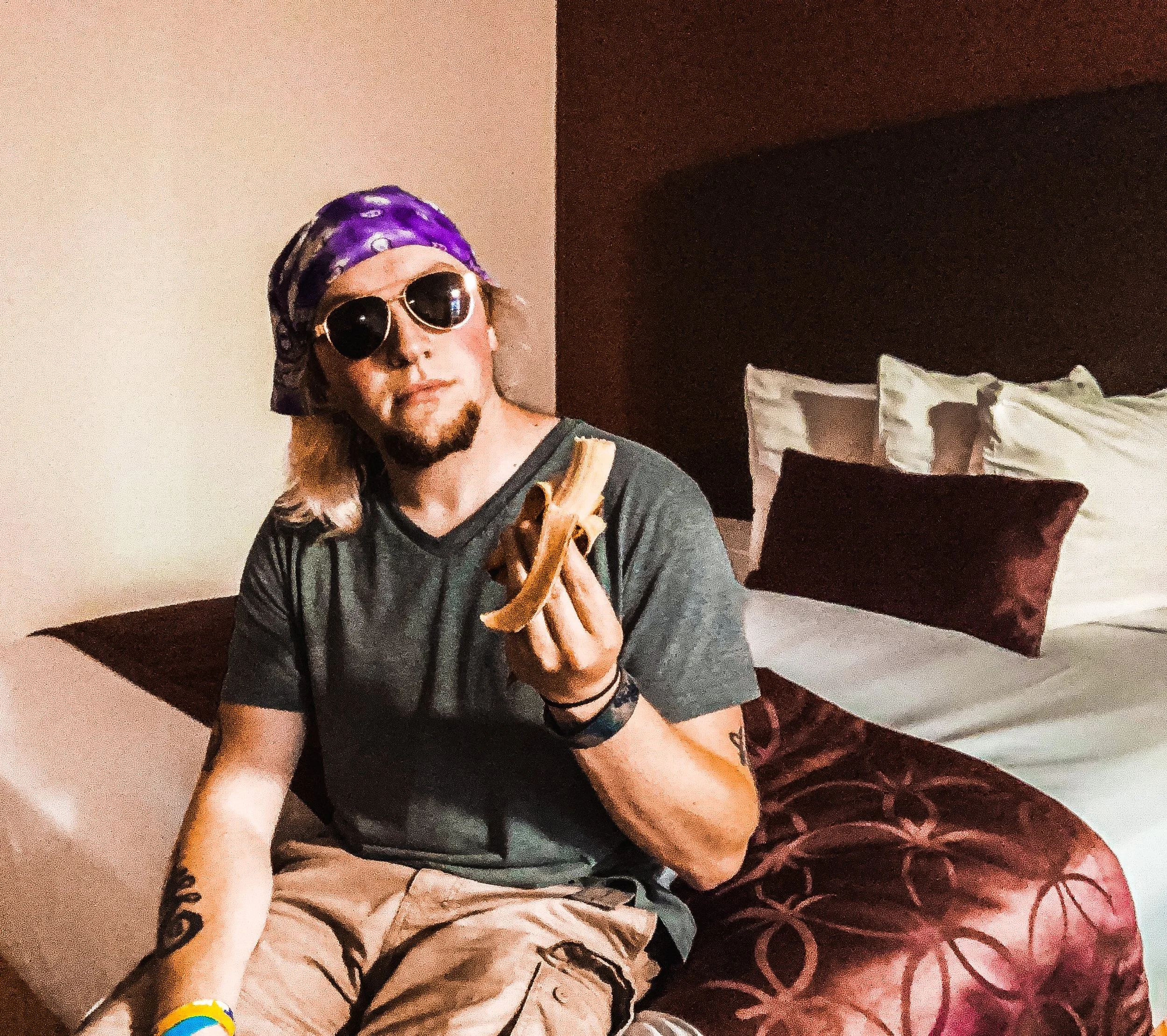 A man with long blonde hair, sunglasses, a purple bandana, and tattoos on his arms sitting on a hotel bed, holding a banana.