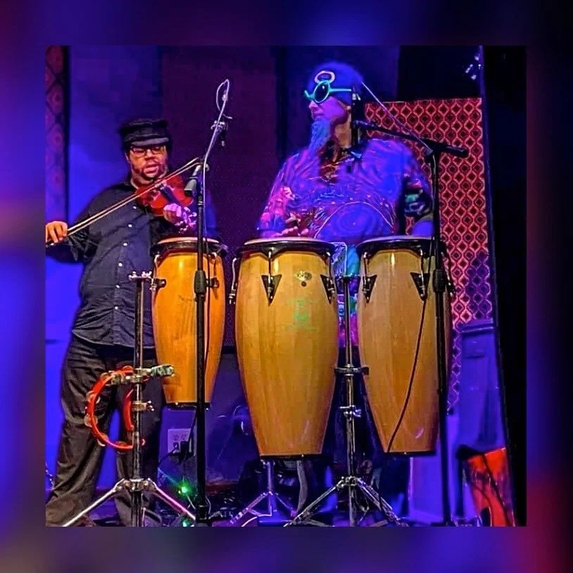 Two musicians performing on stage, one playing a violin and the other playing conga drums, with colorful stage lighting and microphones set up around them.