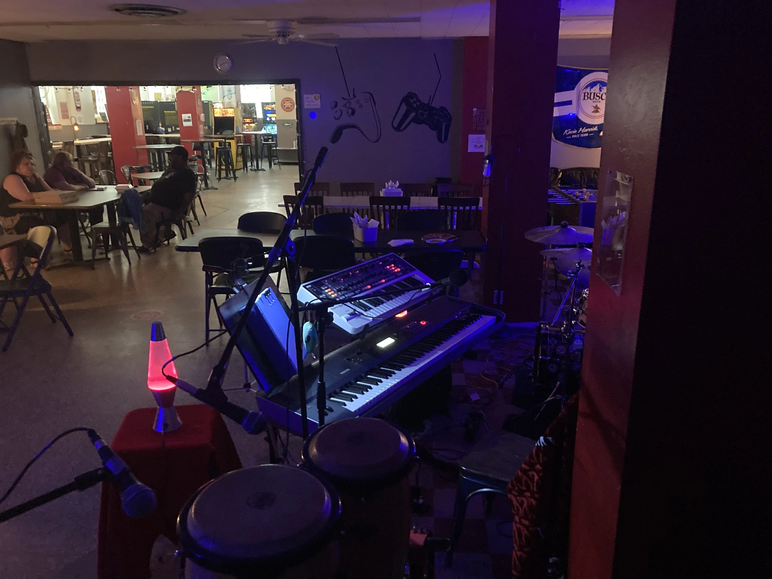 Musical instruments and sound equipment on stage in a dimly lit room, with tables and chairs for an audience and arcade games visible in the background.
