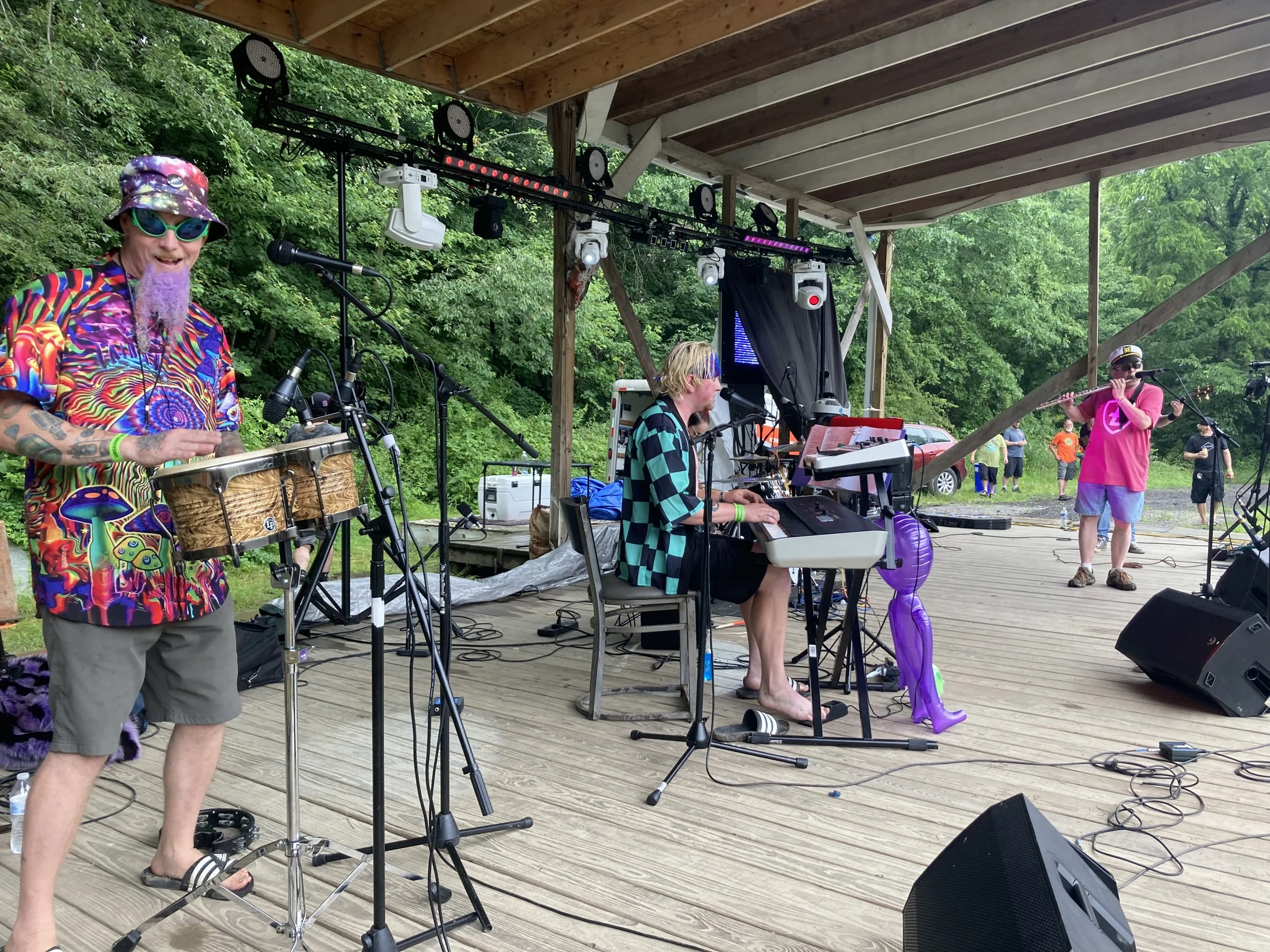Musicians performing on an outdoor stage with a green, wooded background, one playing percussion, another on keyboard, and a third playing the flute, all dressed casually.