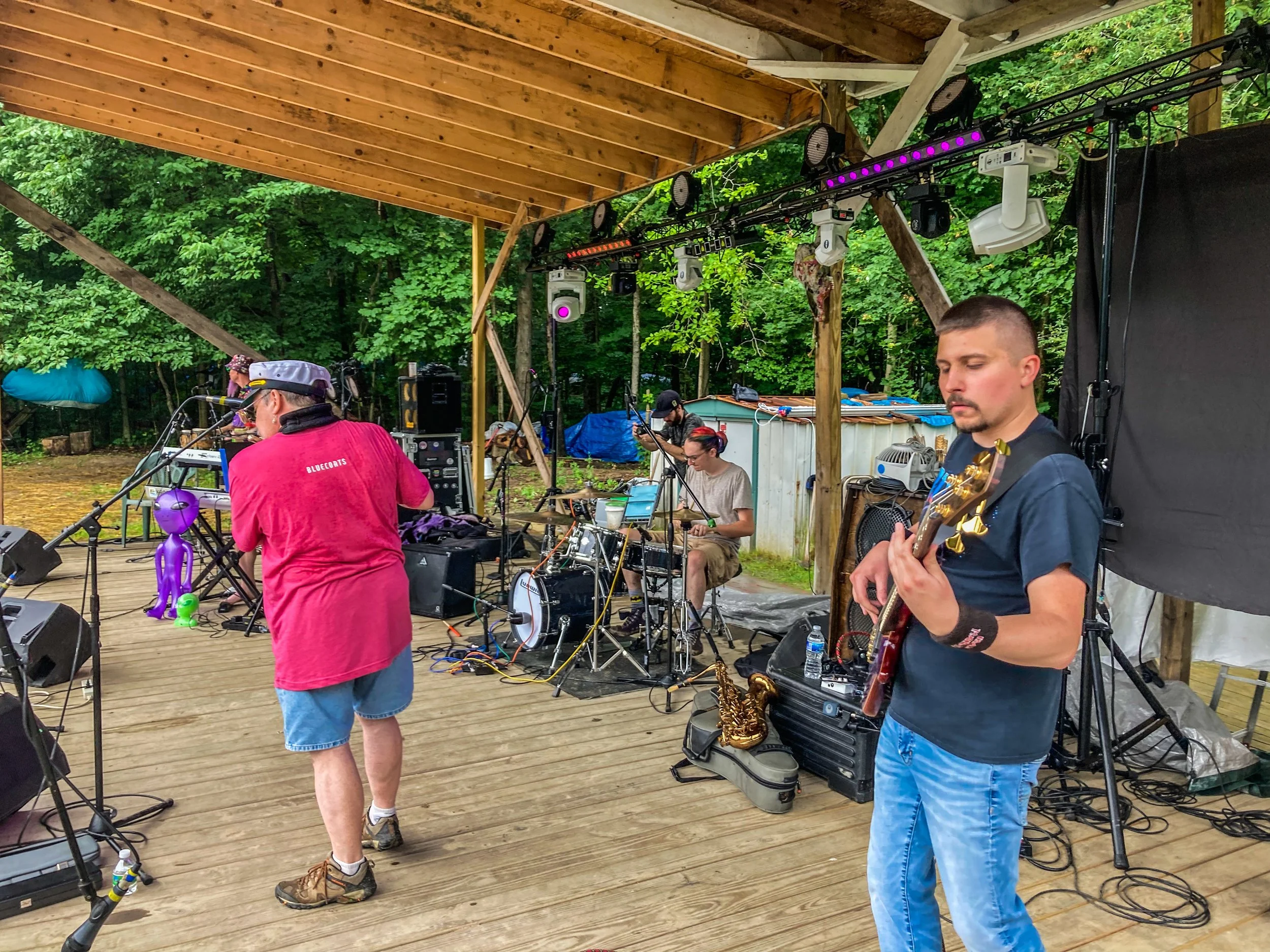 A band performs on a wooden outdoor stage with trees in the background. There are four musicians, and the scene includes musical equipment, stage lighting, and a purple inflatable alien figure.