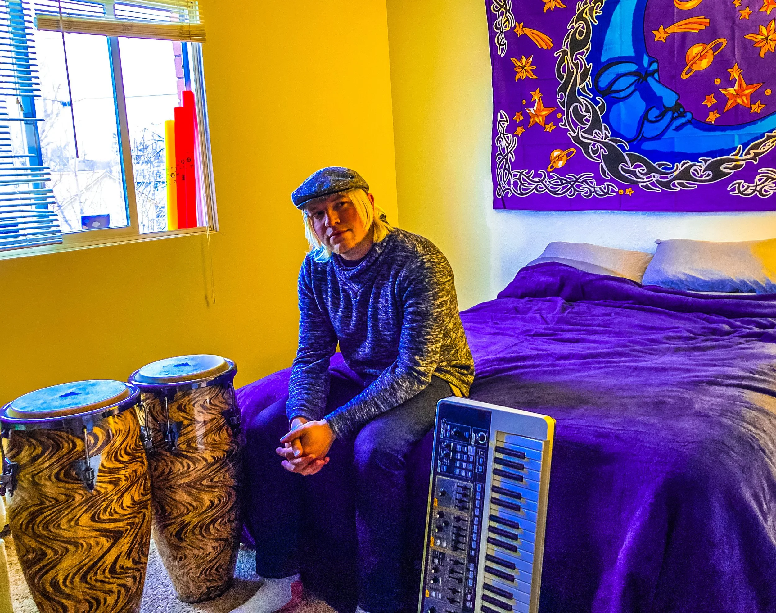 A person with blonde hair wearing a gray hat and gray long-sleeved shirt sitting on a bed in a yellow-walled room, surrounded by musical instruments including two drums, a keyboard, and a keyboard pedal. There's a colorful tapestry with a face and st