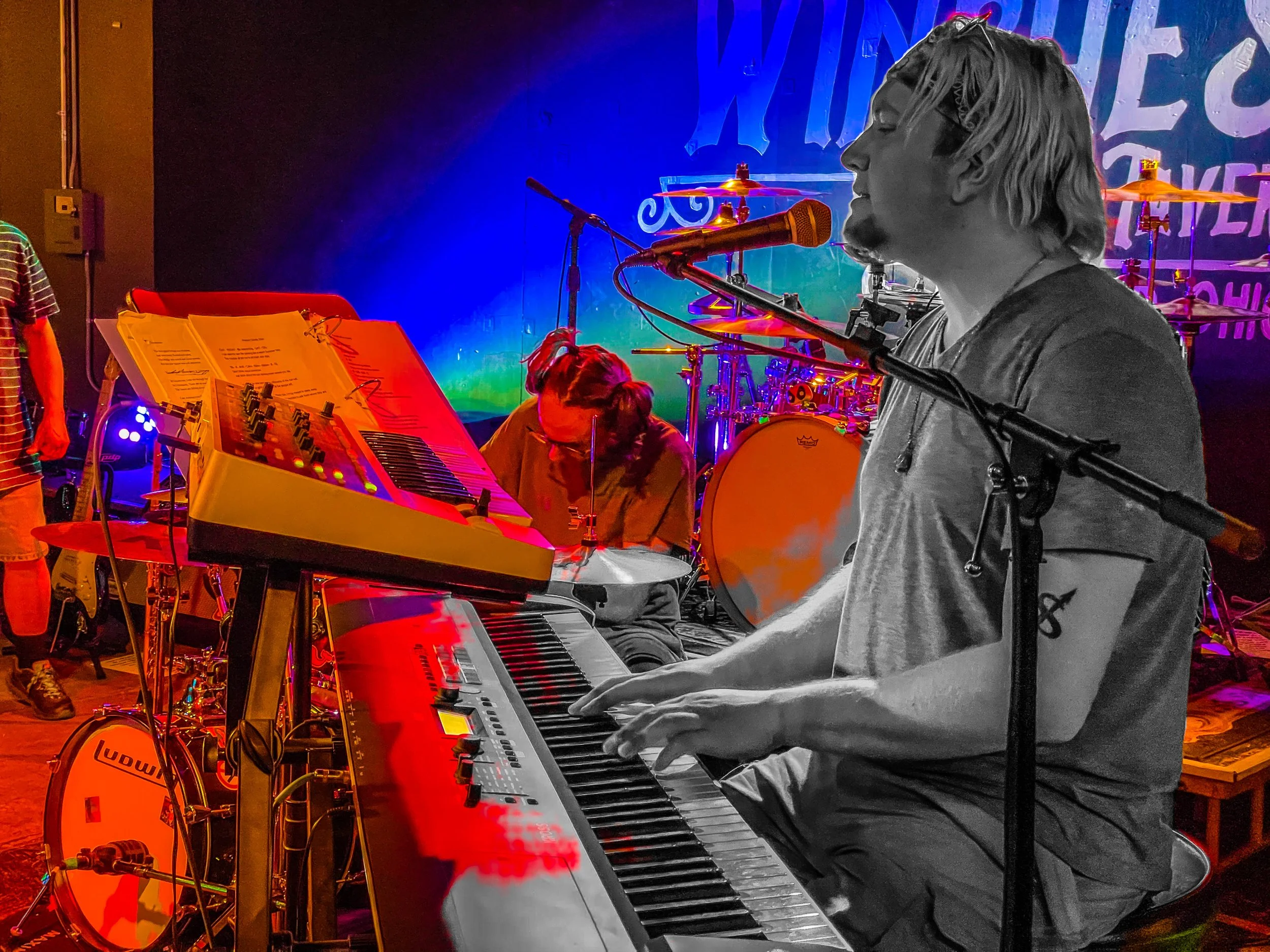 Musicians performing on stage, with a man playing keyboard and singing into a microphone, in front of colorful stage lighting and drums.