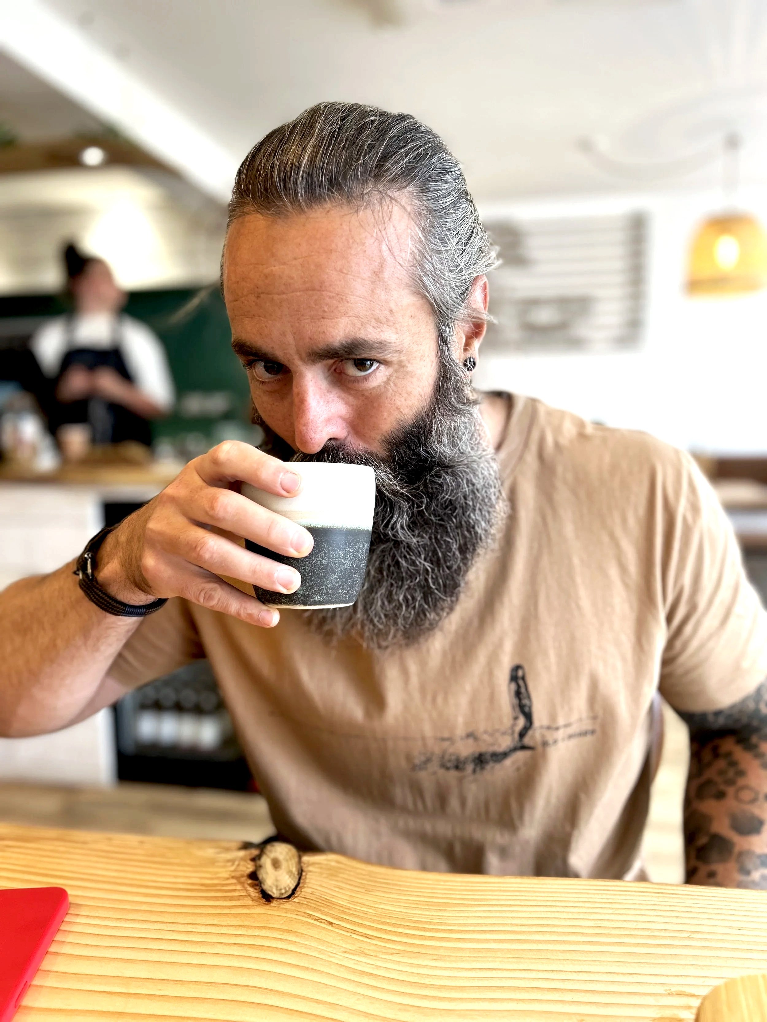 A man with gray hair and a beard holding a glass of dark beer with foam, sitting at a wooden table in a cafe.