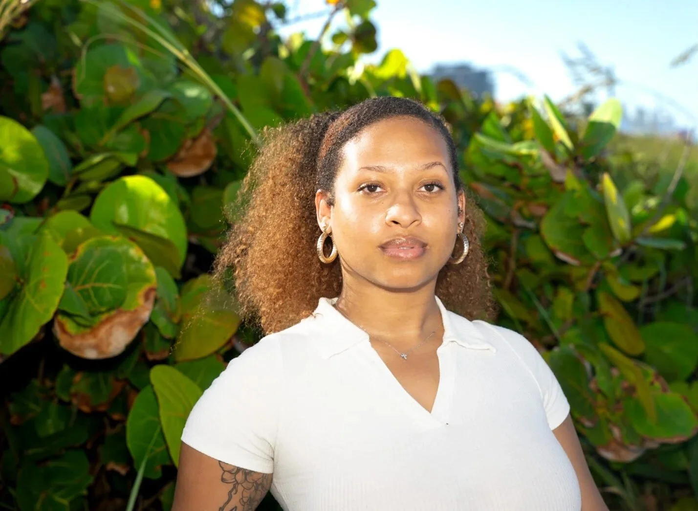 A woman with curly brown hair wearing hoop earrings and a white polo shirt standing outdoors in front of green foliage and some buildings in the distance.