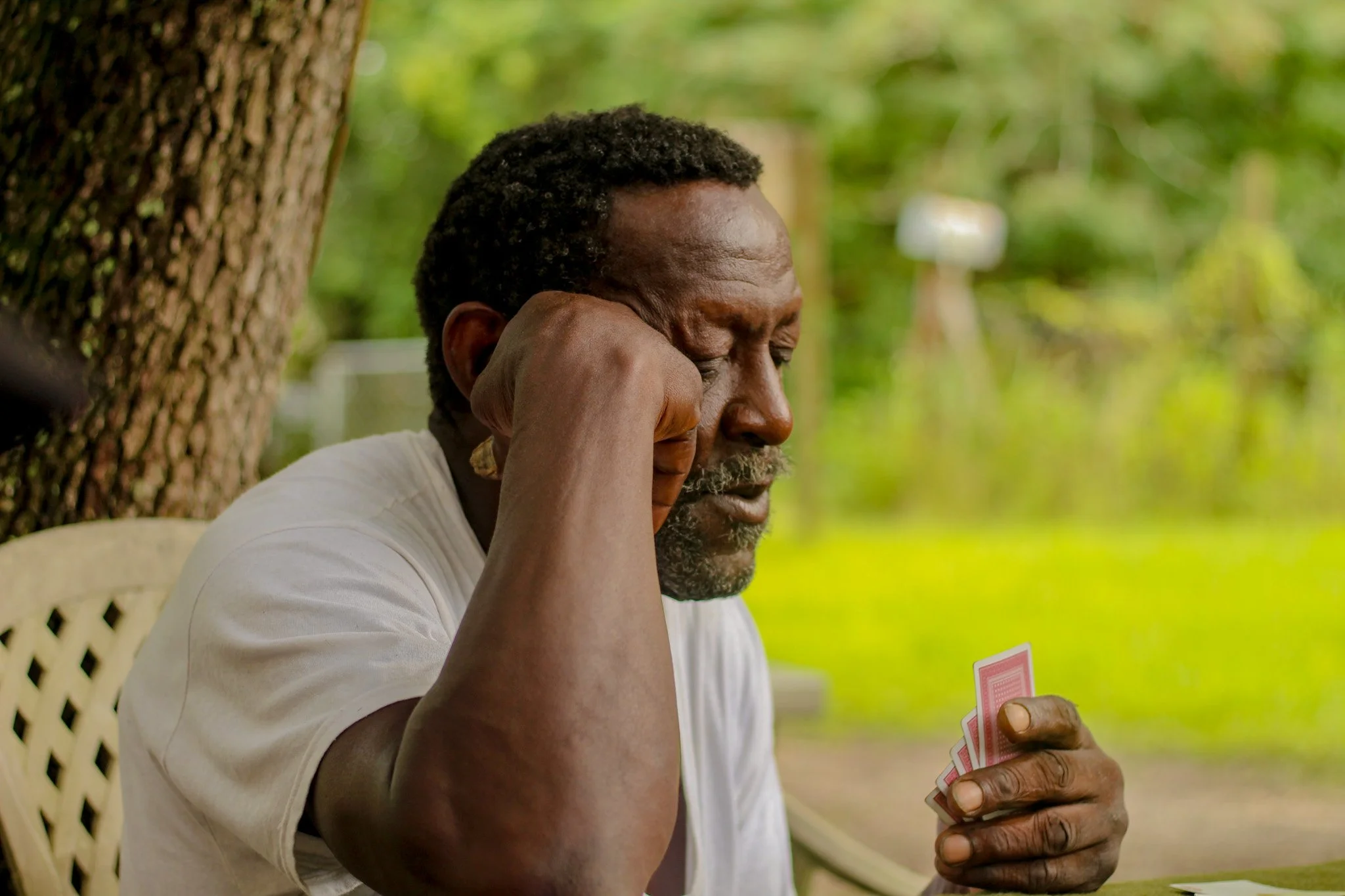 A man sitting outdoors at a table, holding playing cards in his hand, with his face resting on his elbow and eyes closed, in front of a large tree and green foliage.