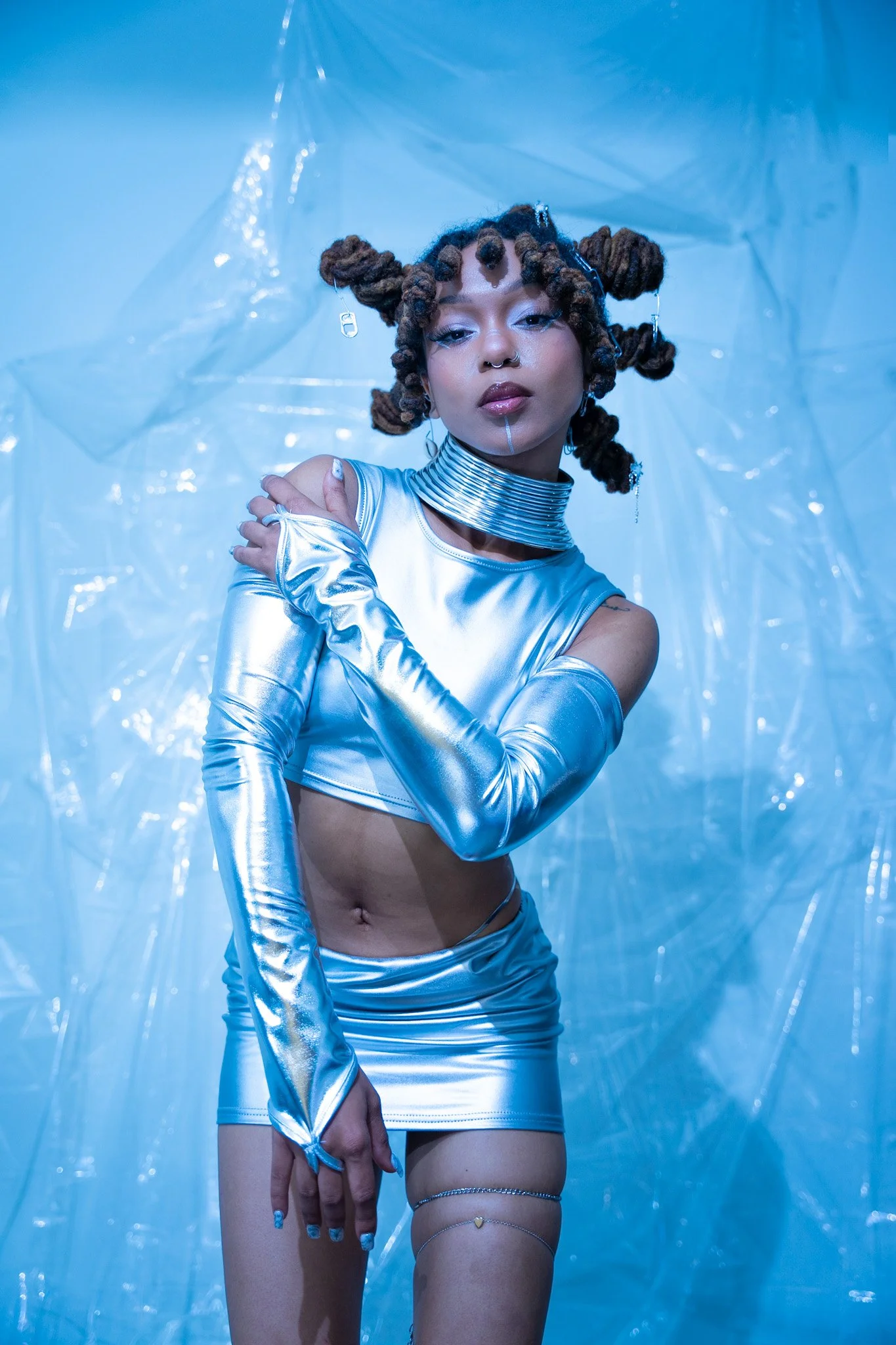 A woman dressed in metallic silver clothing with long matching gloves and layered jewelry, posing against a light blue background with plastic sheeting.