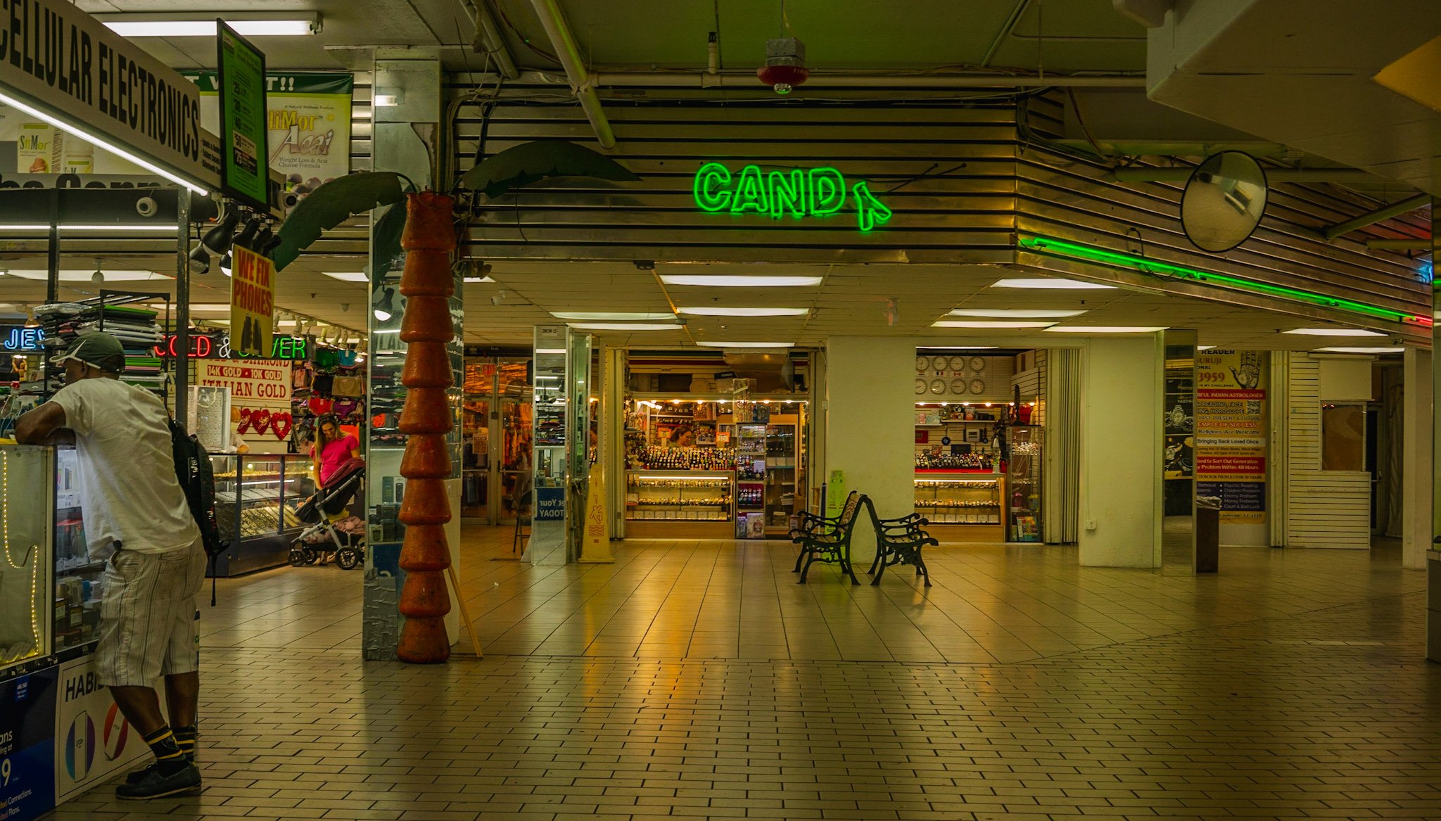 An indoor marketplace or shopping area with a neon green sign that says 'CAND' and an artificial palm tree. There are several small shops with illuminated displays, a few people shopping, and benches in the center.