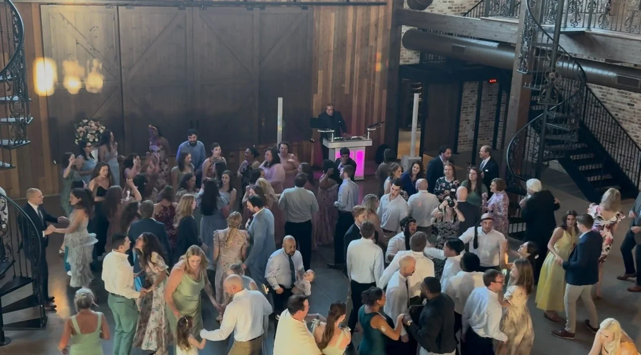 People dancing and socializing at a wedding reception in a spacious indoor venue with wooden walls, a staircase with ornate black railing, and a DJ booth with pink lighting.