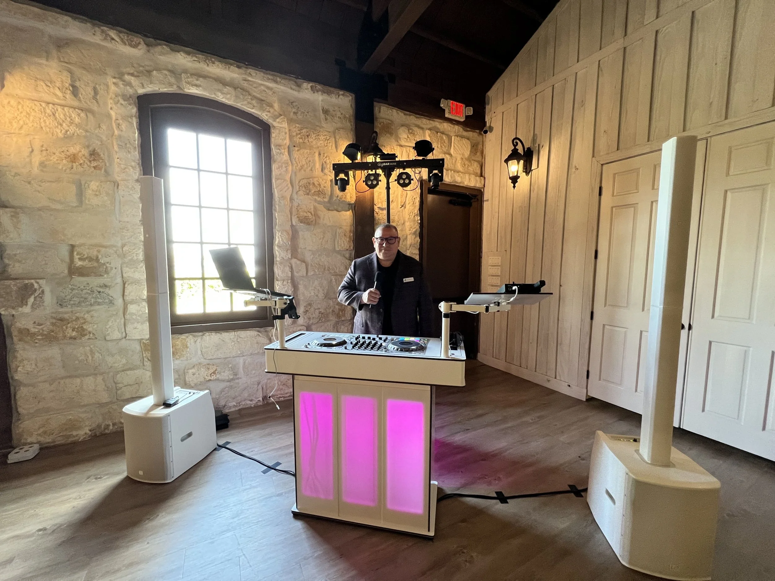 A DJ stands behind a DJ booth with colorful lights in a rustic room with stone walls and wooden doors, large windows, and vintage wall sconce lighting.
