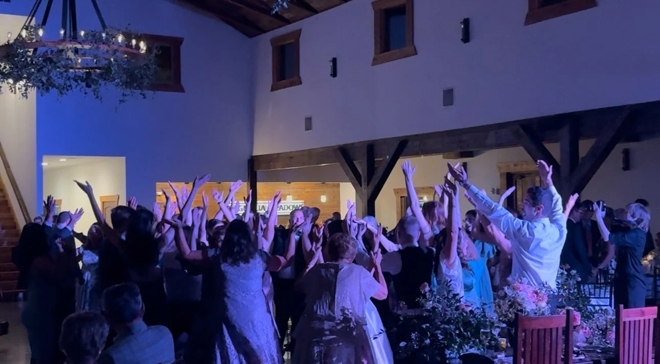 People dancing and celebrating at a wedding reception with colorful lighting and floral decorations.