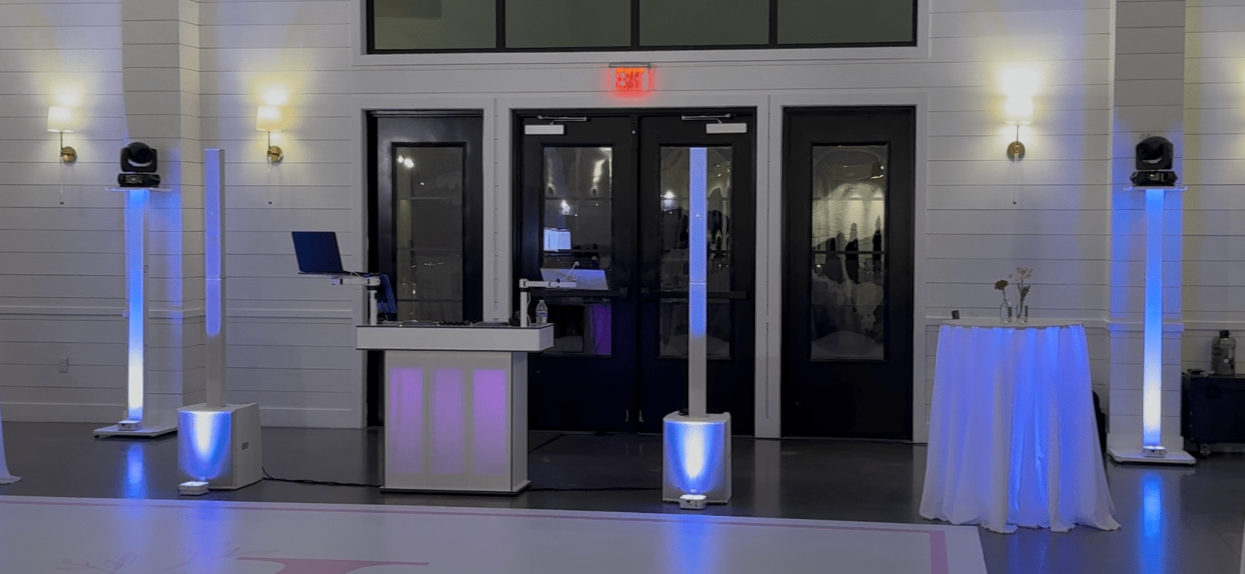 Event setup with DJ equipment, tall speakers with blue lights, a small round table with flowers, and a white tablecloth, in a room with white walls and black double doors.