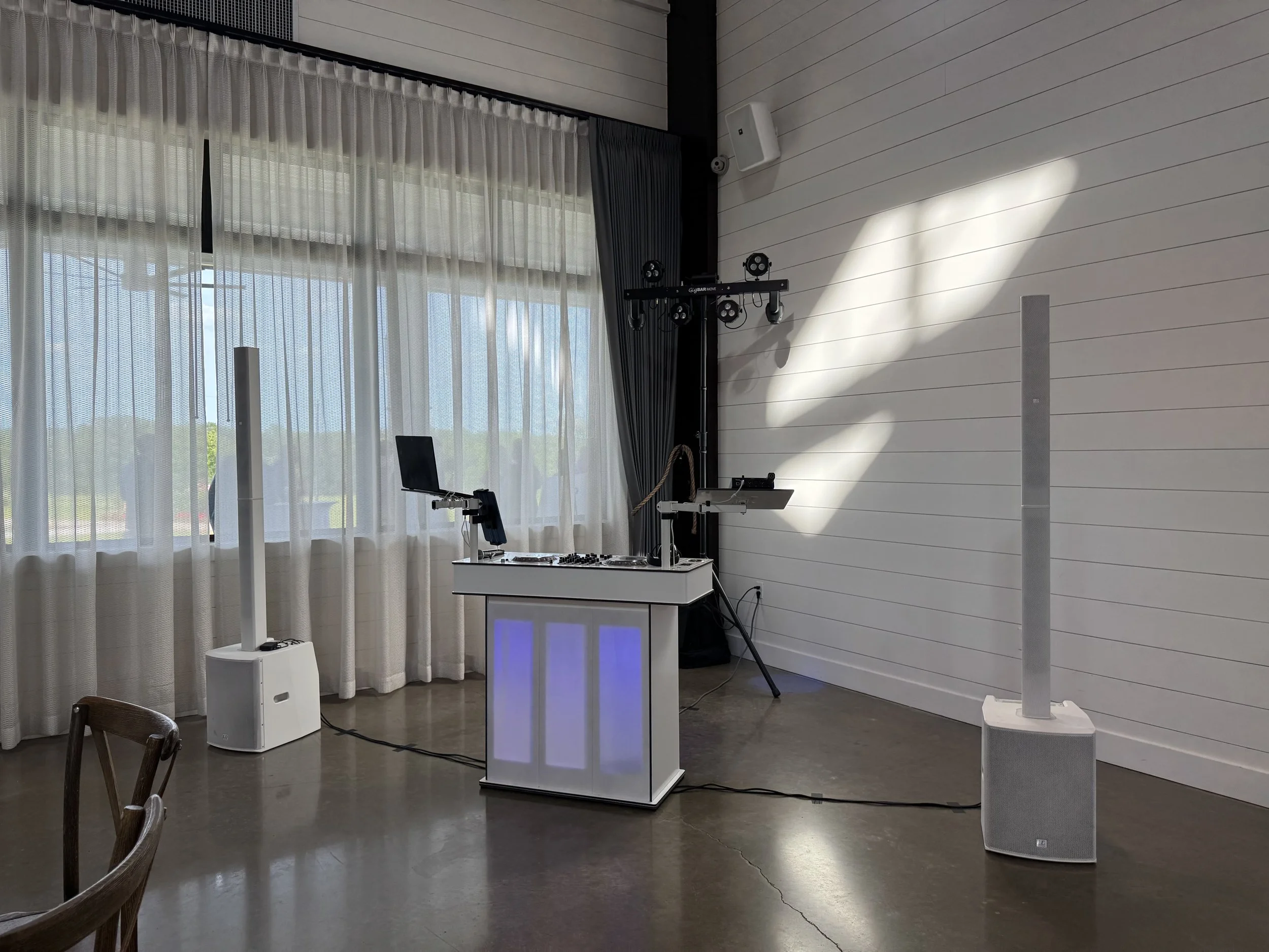DJ booth setup with speakers, lighting, and a laptop in a room with large windows and white walls.