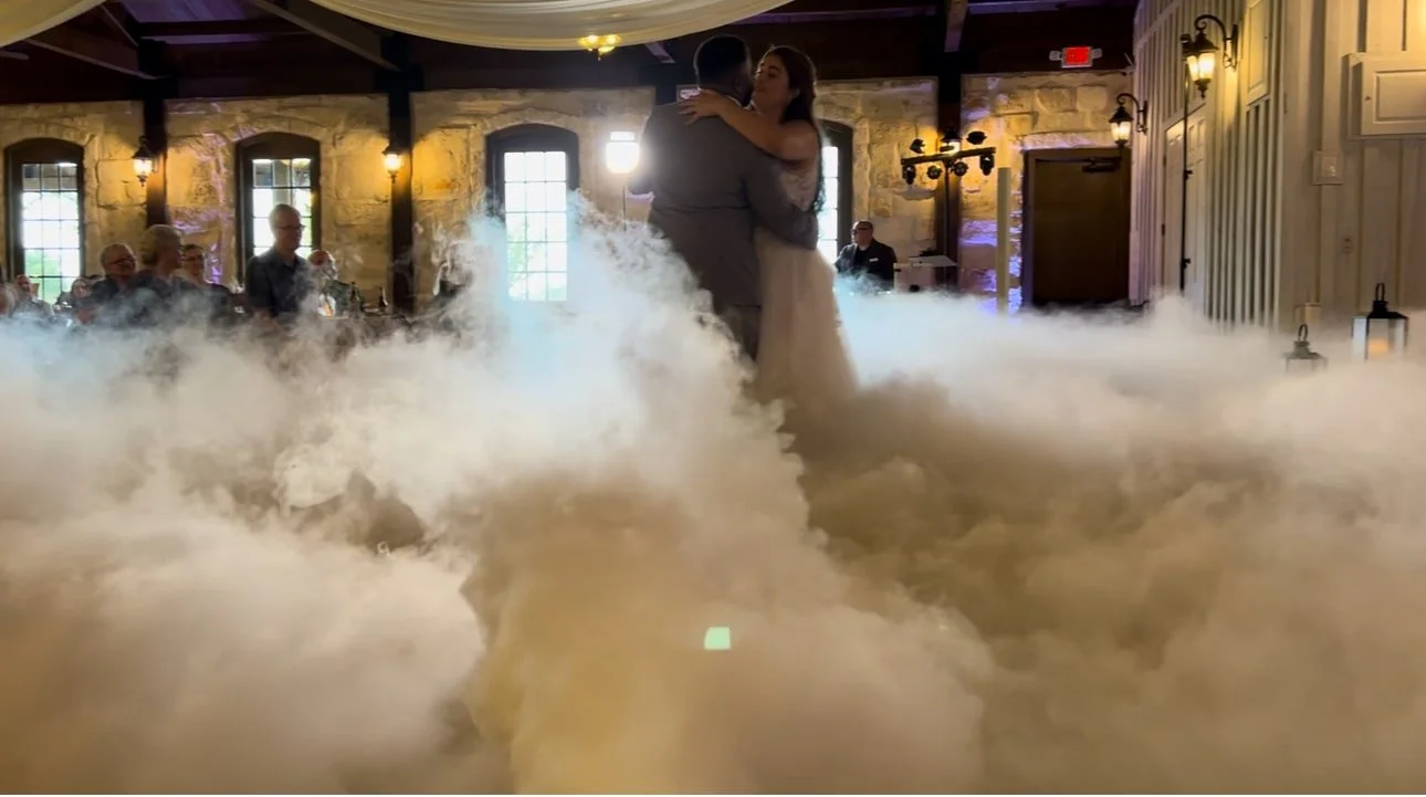 A bride and groom sharing a first dance in a wedding reception hall with a fog effect and guests watching.