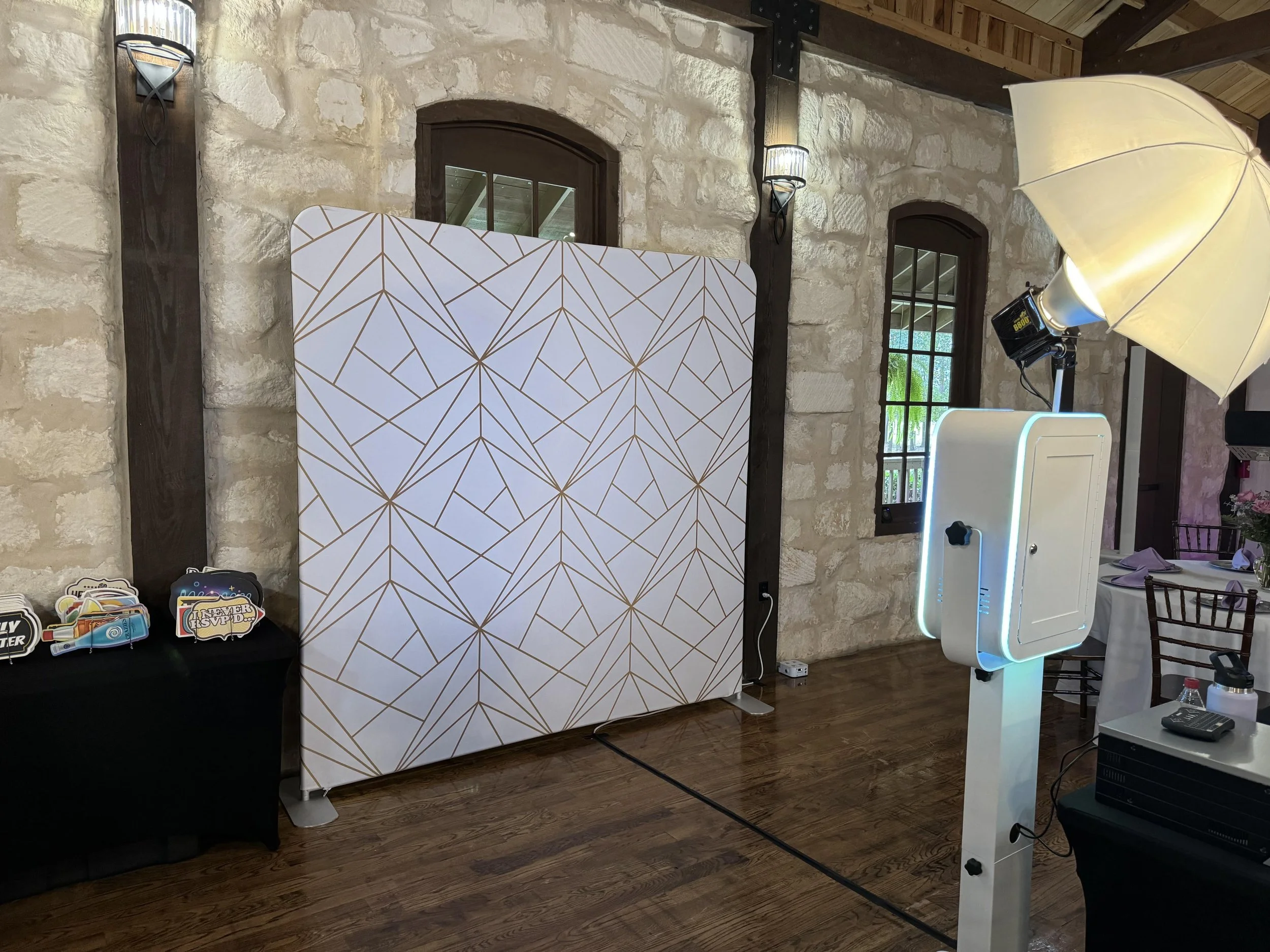Photo booth setup with a geometric patterned white backdrop, photo props on a black table, and a professional lighting and camera system in a rustic room with stone walls and windows.