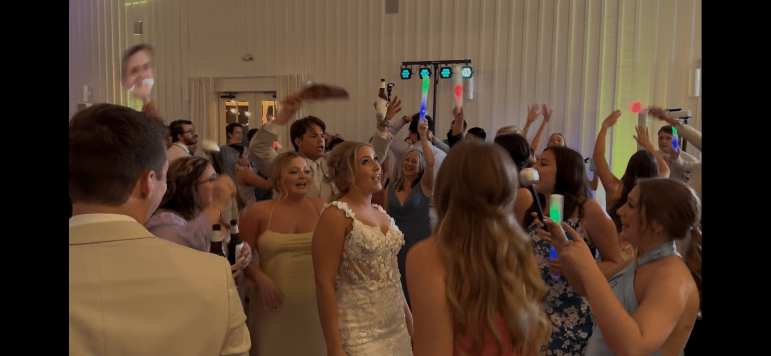 People dancing and celebrating at a wedding reception with colorful lighting and a lively atmosphere.