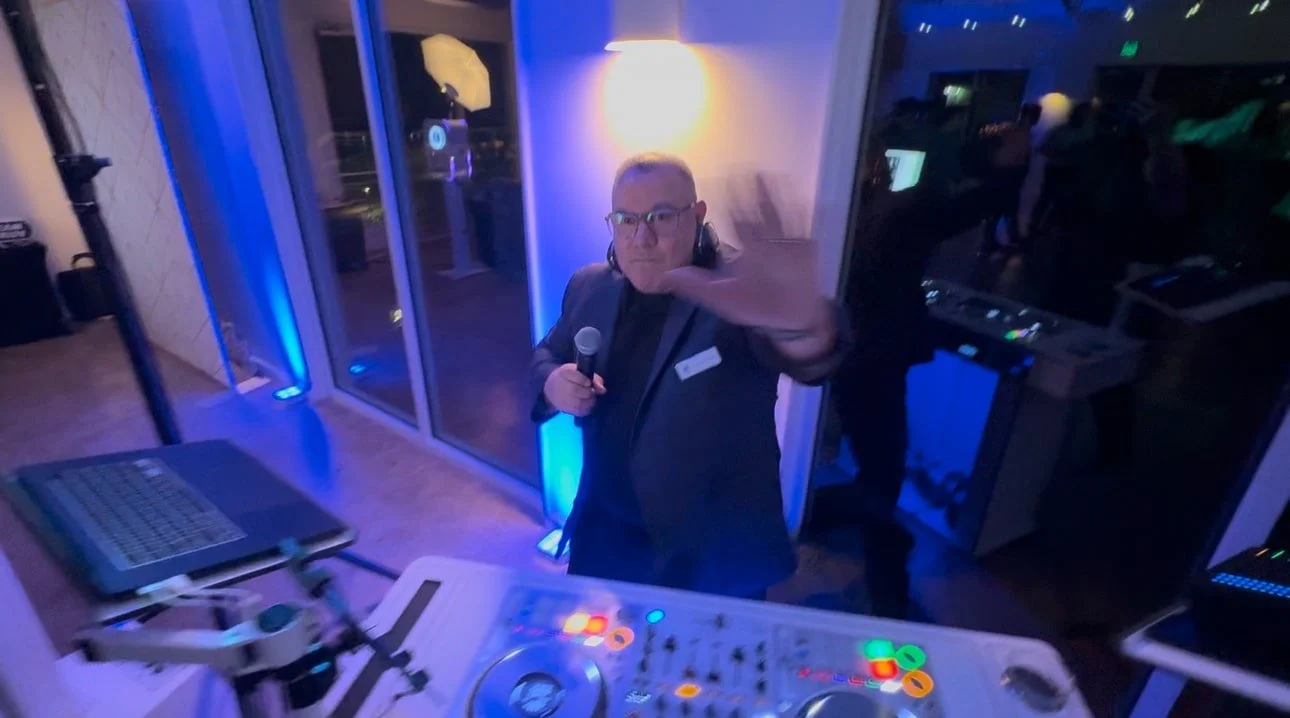 A DJ holding a microphone and waving at the camera at a party or event, with DJ equipment and a laptop in front of him, illuminated by colorful lighting.
