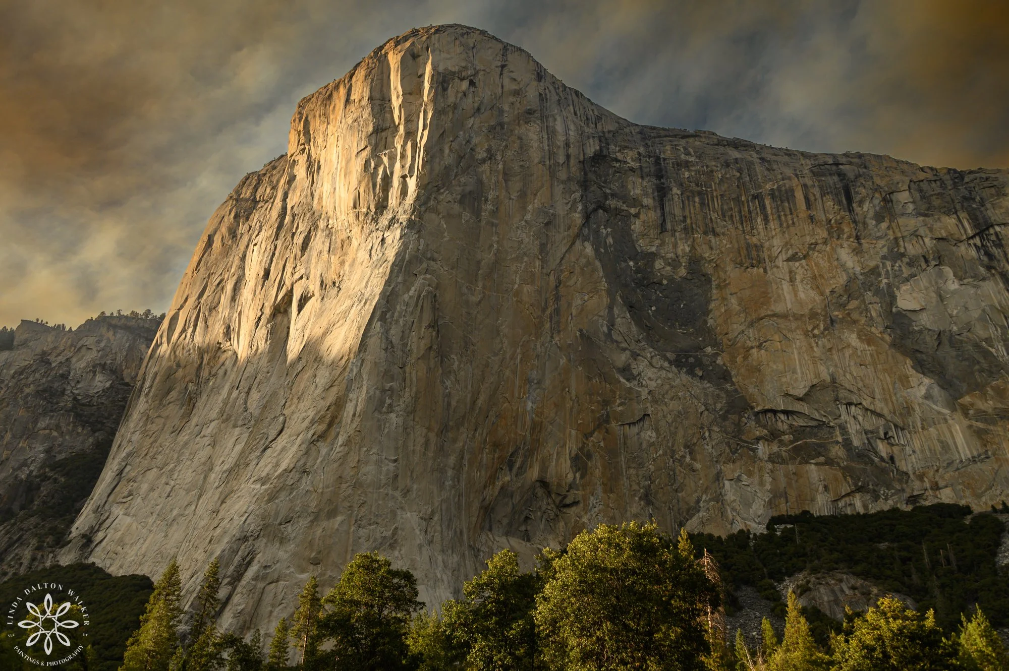 The sheer granite wall of El Capitan illuminated by warm orange sunset light through wildfire haze in Yosemite Valley.