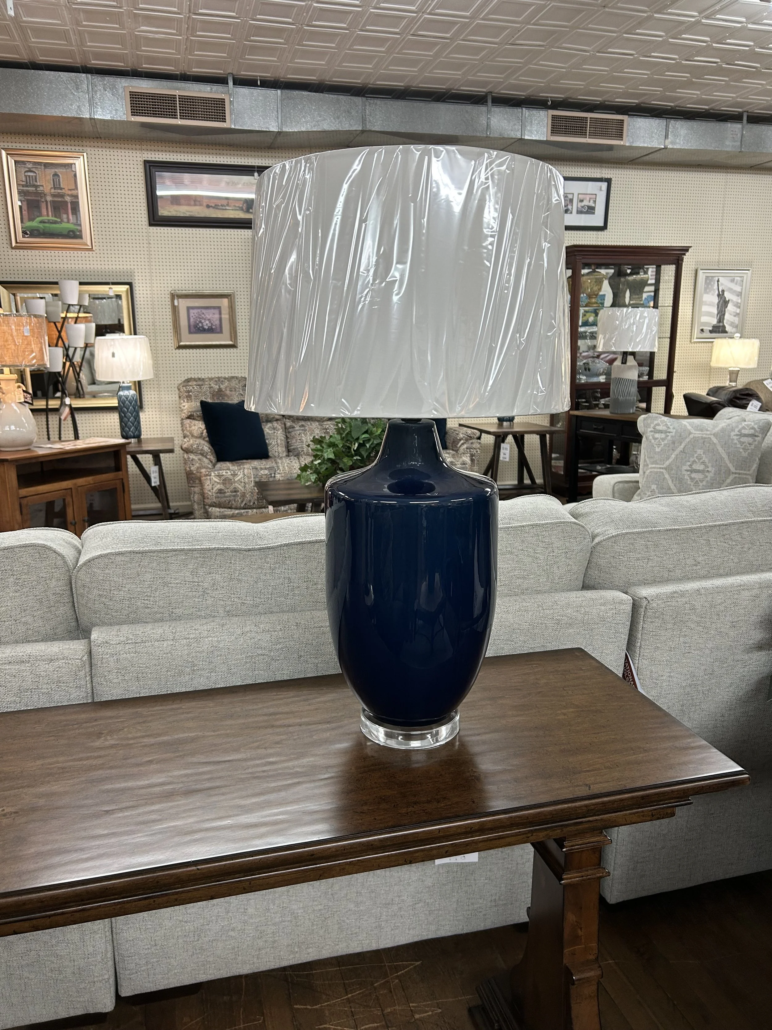 A large blue ceramic table lamp with a white fabric lampshade wrapped in clear plastic, placed on a wooden table in a furniture store.