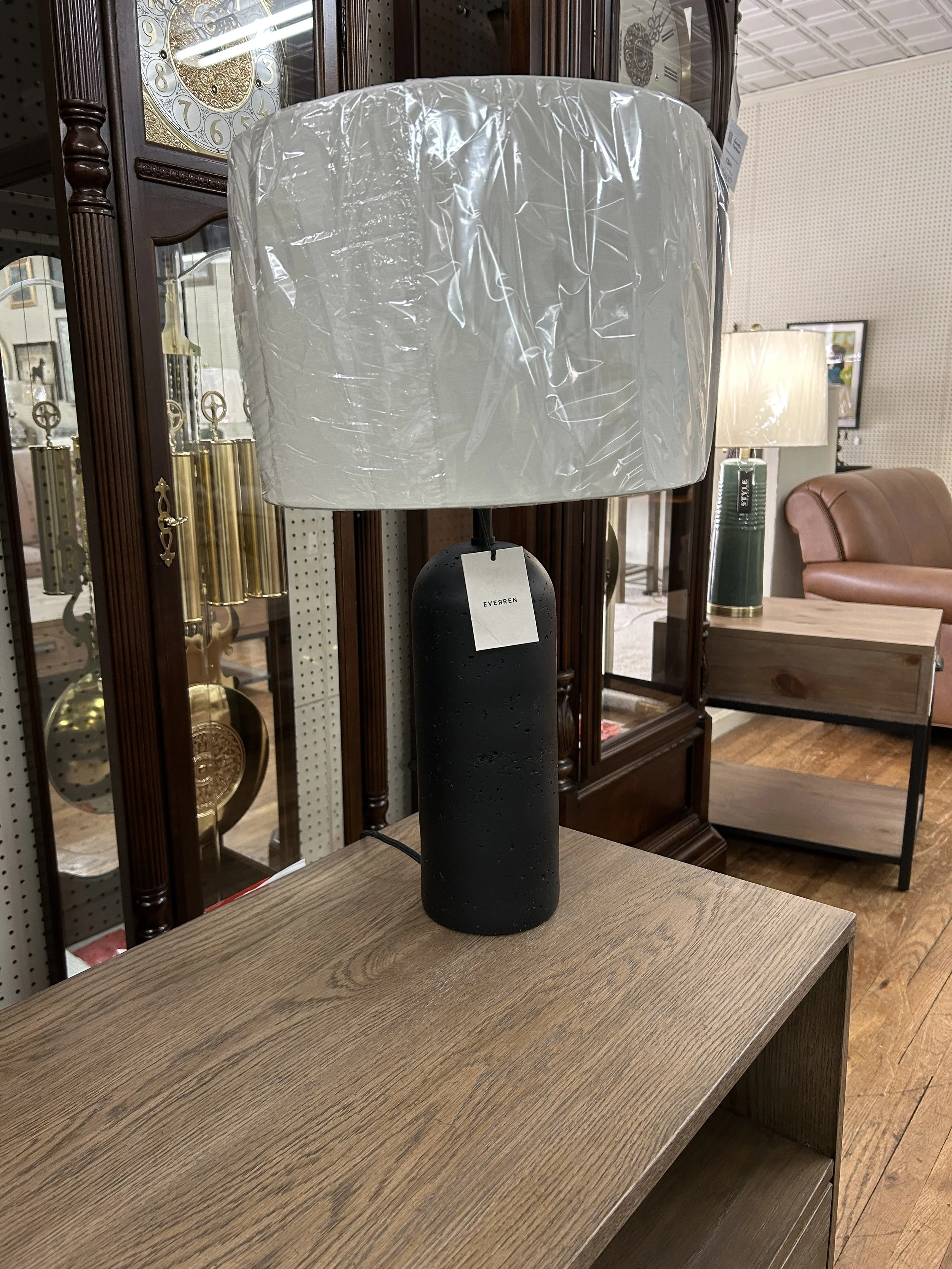 A black table lamp with a large white plastic wrap-covered lampshade on a wooden surface in a furniture store.